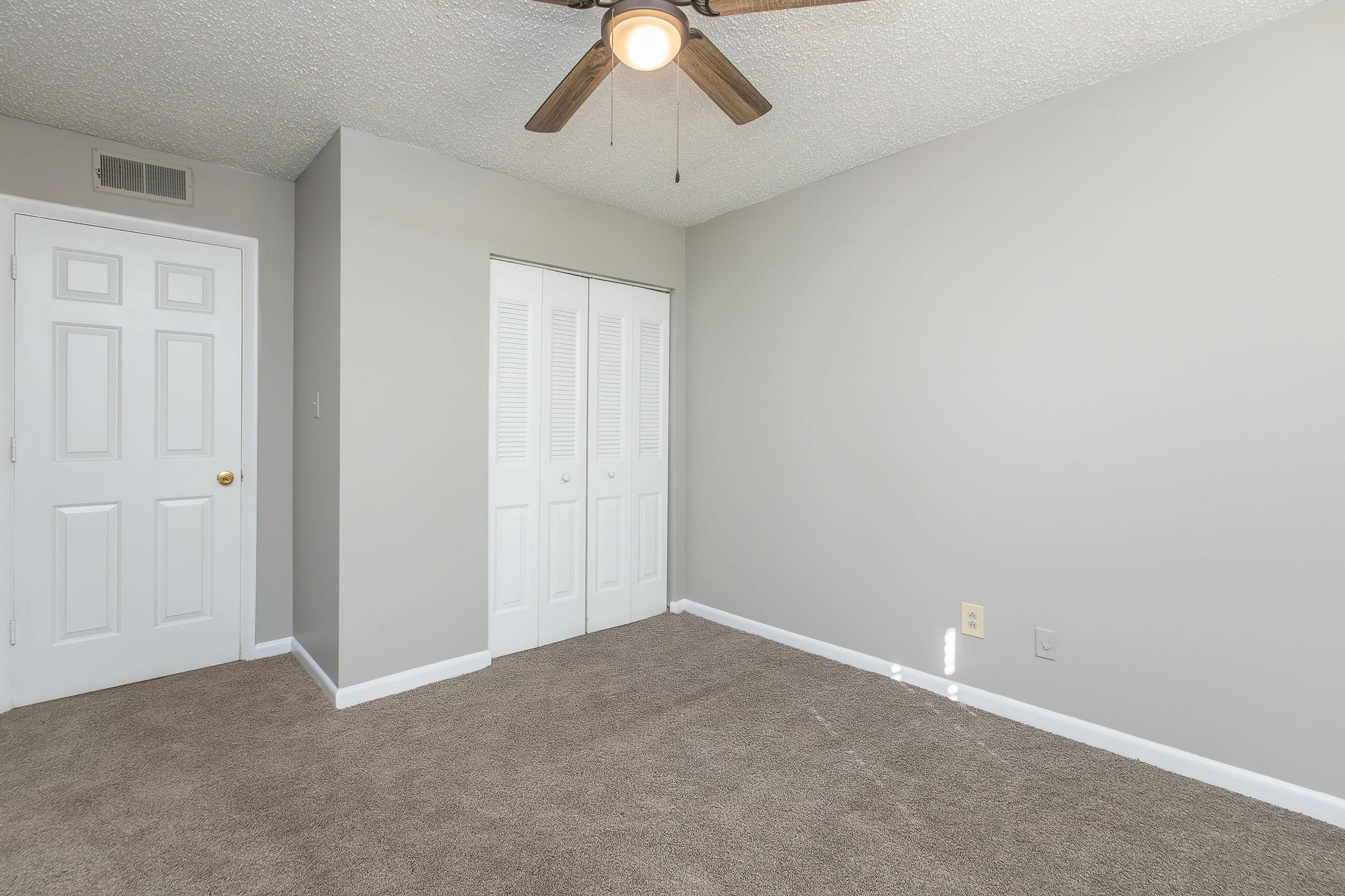 A light-filled empty bedroom with beige carpet and a ceiling fan. The walls are painted light gray, and there are double white closet doors. The room has no furniture, providing a clean, spacious look. A small portion of sunlight is visible on the floor.