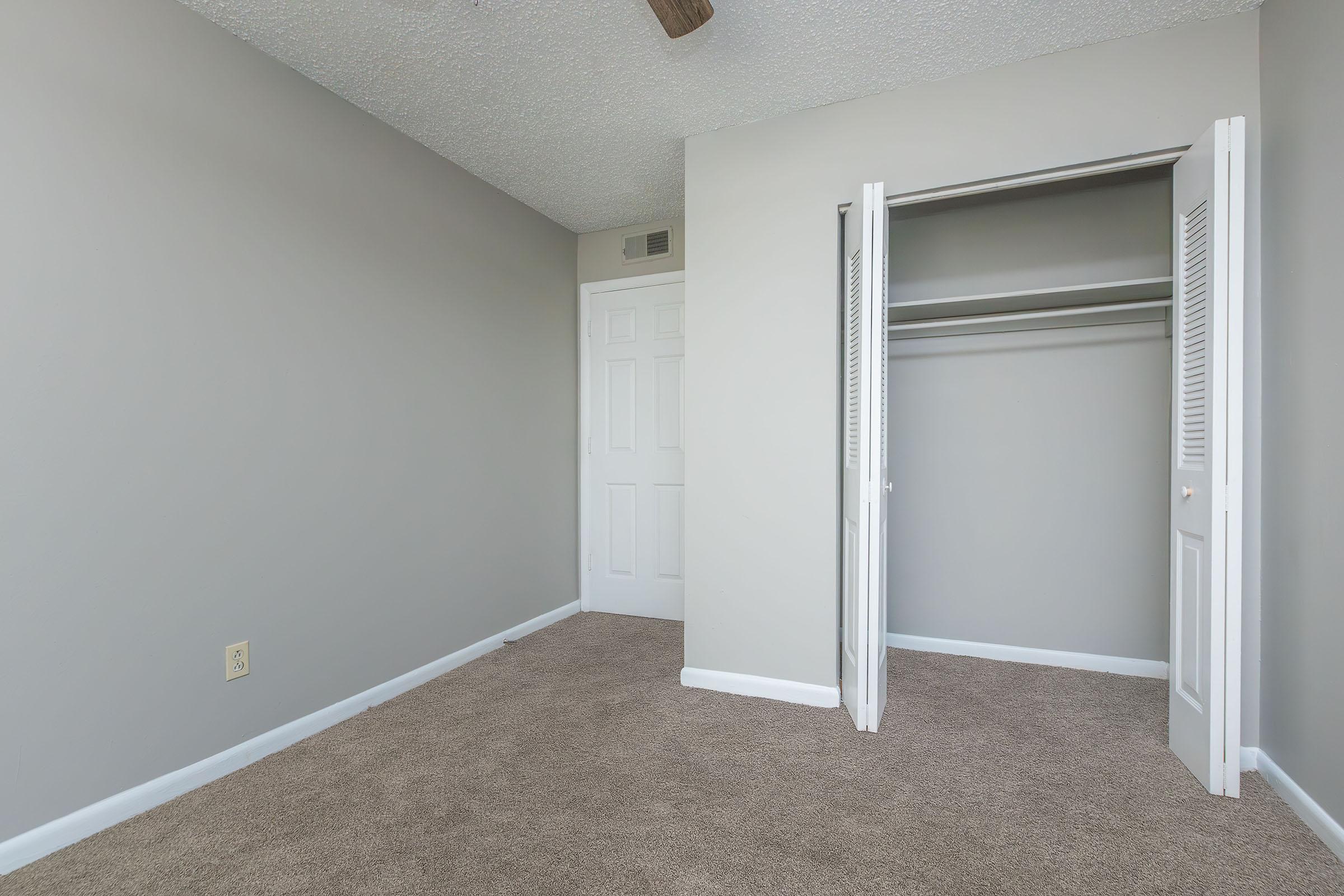 A spacious, empty room featuring light gray walls and a ceiling fan. The floor is carpeted in a neutral color. There is a closed door leading to a closet on the right, which has white louvered doors. Natural light brightens the space, creating a clean and inviting atmosphere.
