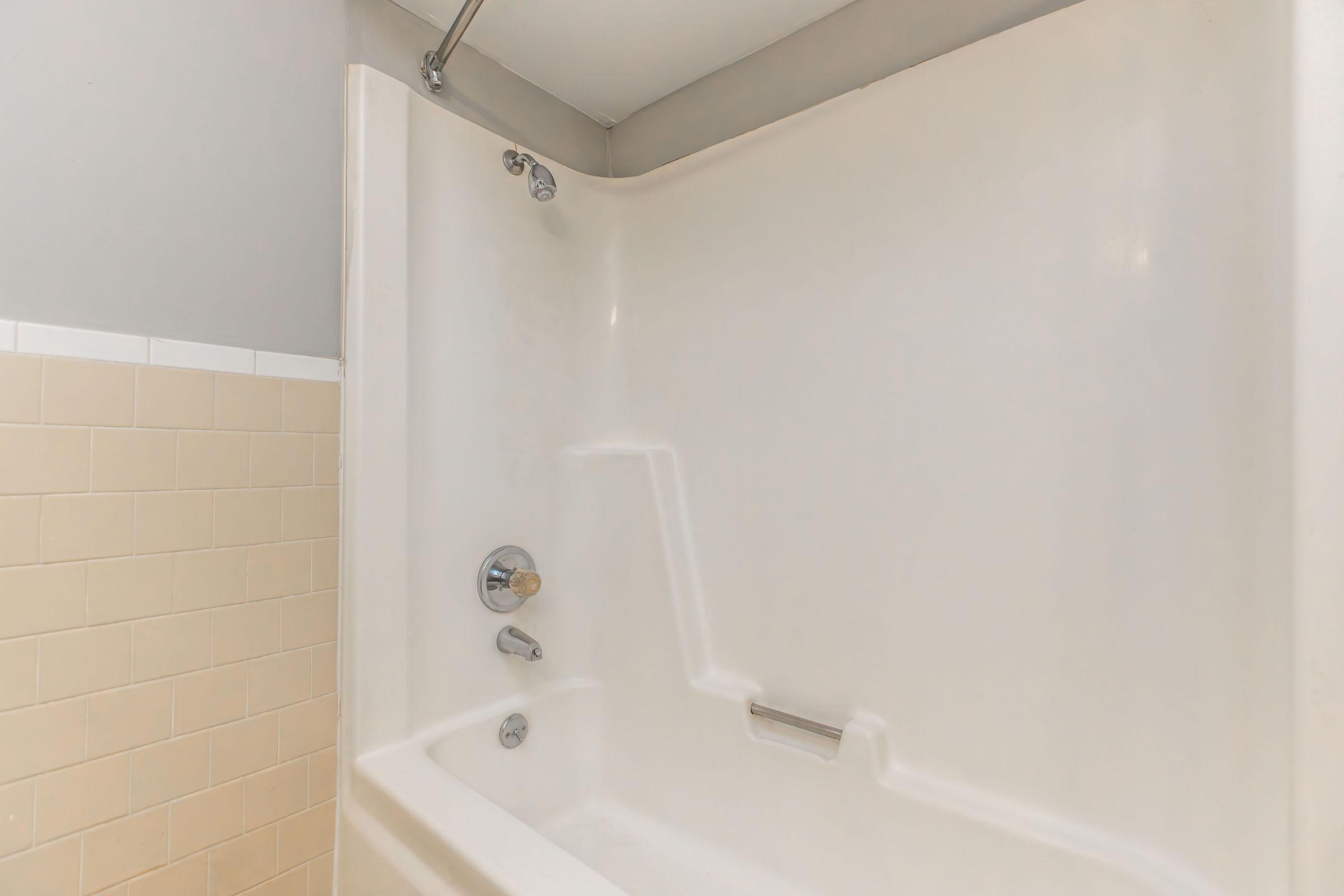 A clean, empty shower stall featuring a white tub and tiled wall in a light beige color. The showerhead and faucet are visible on the wall, and the space is well-lit, creating a fresh and airy atmosphere.