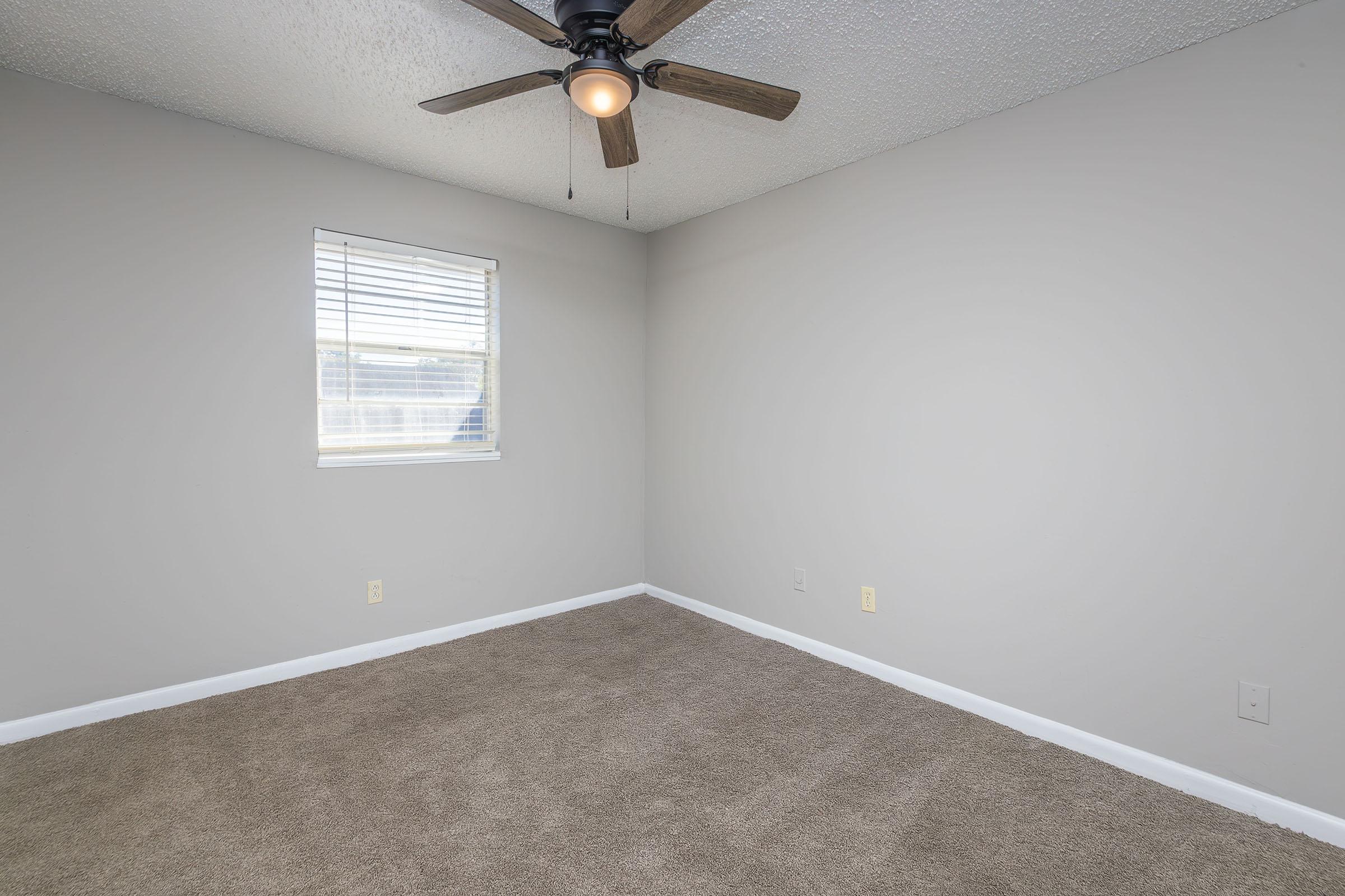 A clean, empty room with light gray walls and beige carpet. There is a window with blinds on the left side and a ceiling fan with wooden blades in the center. The room has no furniture, creating a spacious and blank canvas feel.
