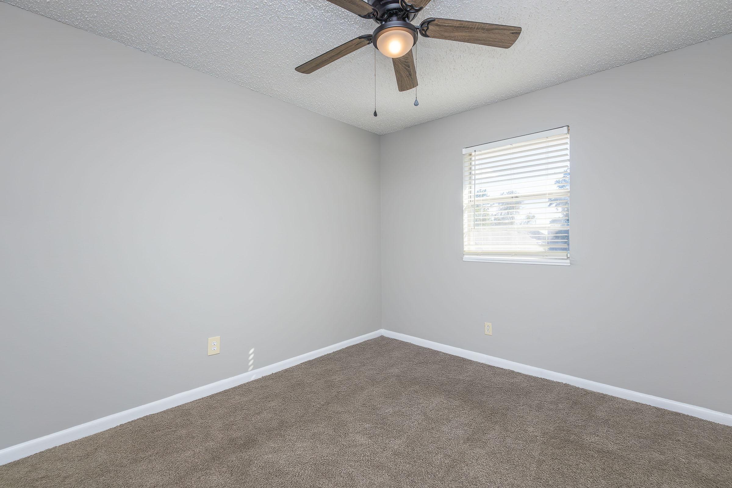 Empty room with light gray walls and a textured ceiling. A ceiling fan with wooden blades is installed, and there is a window with horizontal blinds letting in natural light. The floor is covered with light brown carpet.