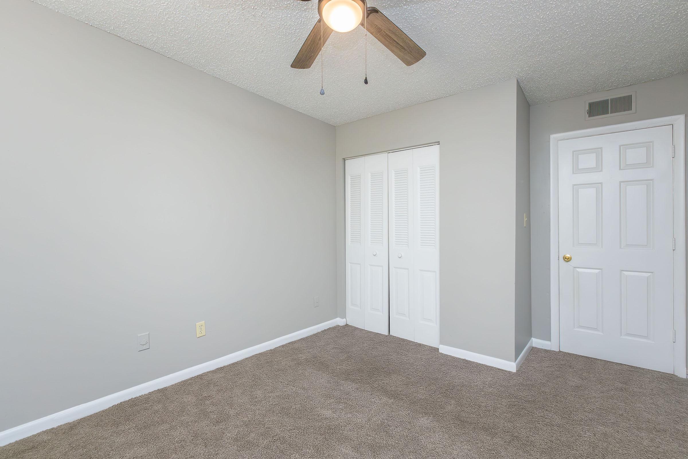 A spacious, empty bedroom featuring light gray walls and carpeted flooring. There is a ceiling fan with wooden blades, a double door closet, and a white door leading to another room. The room is well-lit, offering a neutral and inviting atmosphere.