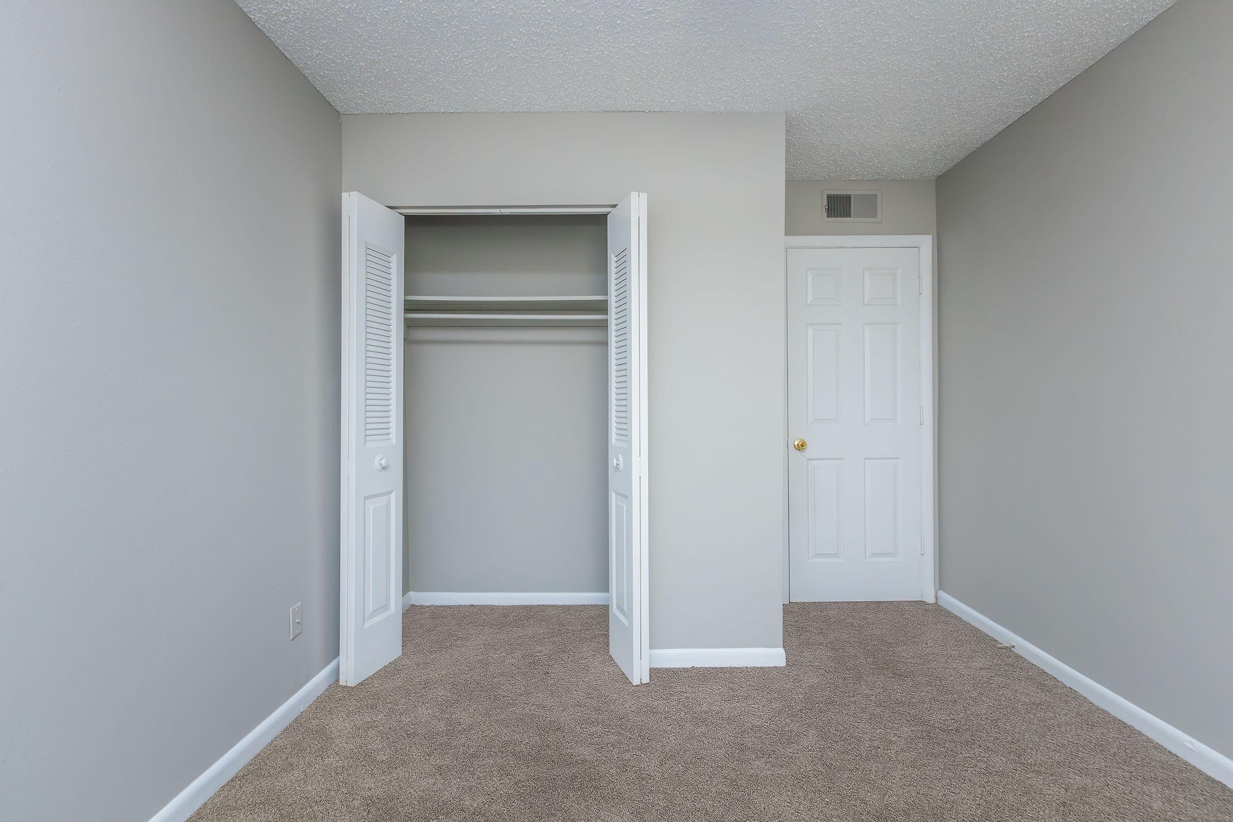 A minimalistic bedroom with light gray walls and beige carpet. The room features an open closet with double doors and a white door leading to another space. Natural light illuminates the area, creating a neutral and inviting atmosphere.