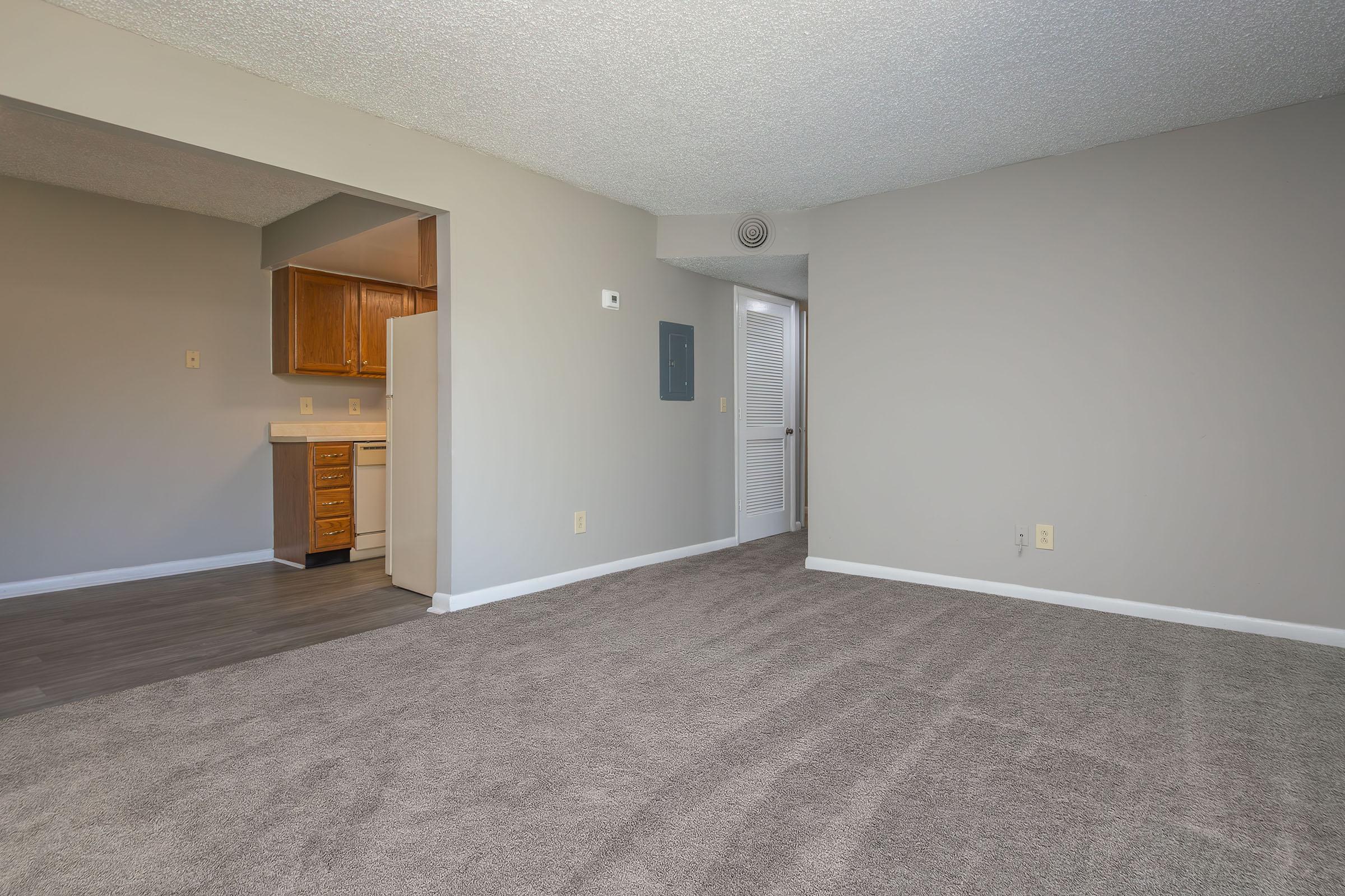 Interior view of a vacant room with gray carpet and light gray walls. To the left, an open doorway leads to a kitchen with wooden cabinets. A white door on the right leads to another room. The ceiling has a textured finish, and there’s ample natural light coming in from the left side of the image.
