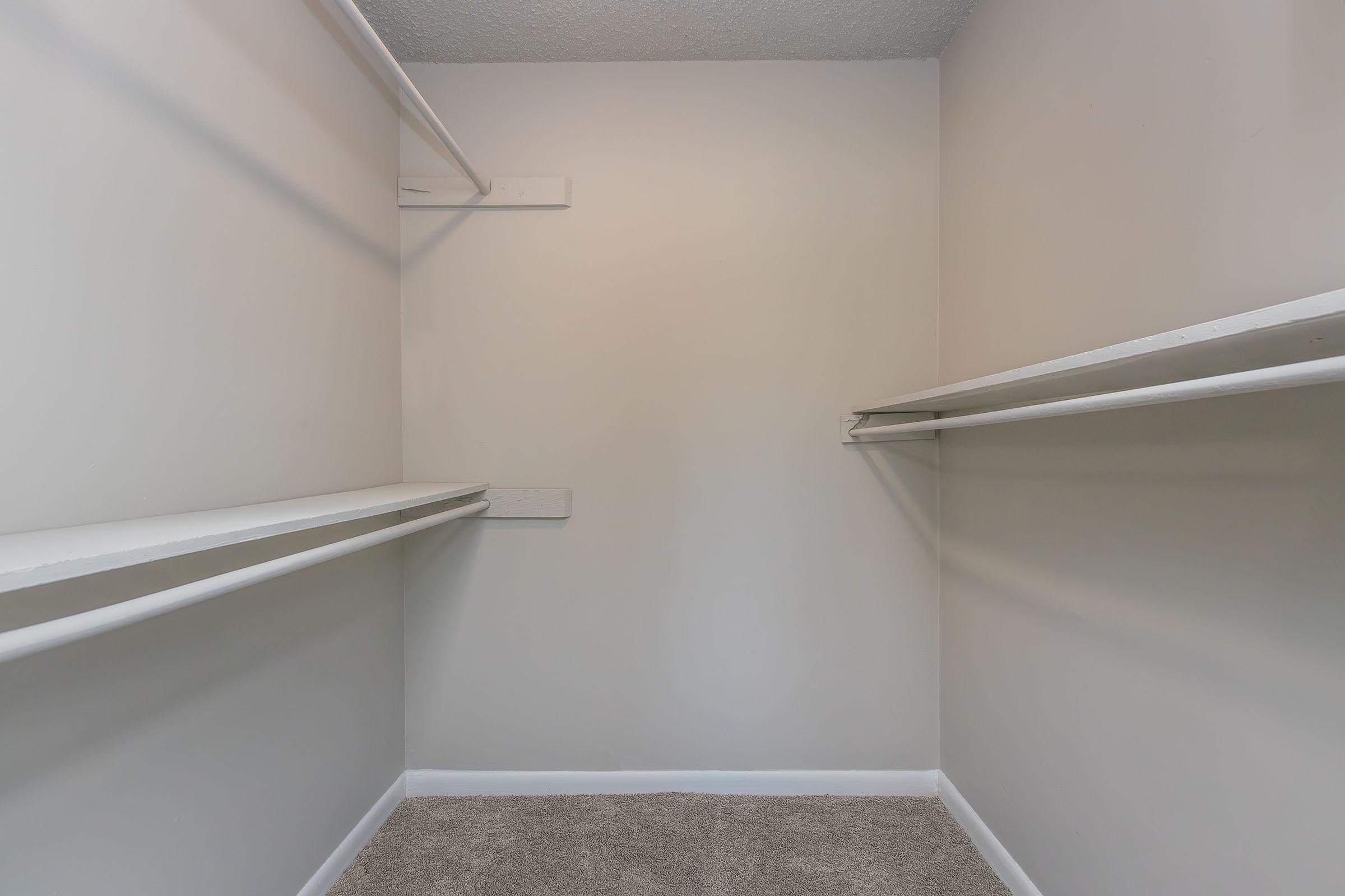 Empty closet with light beige walls, featuring two white shelves installed on opposite sides. The floor is covered with a light-colored carpet, and the ceiling is visible above, showing a plain and unadorned surface.