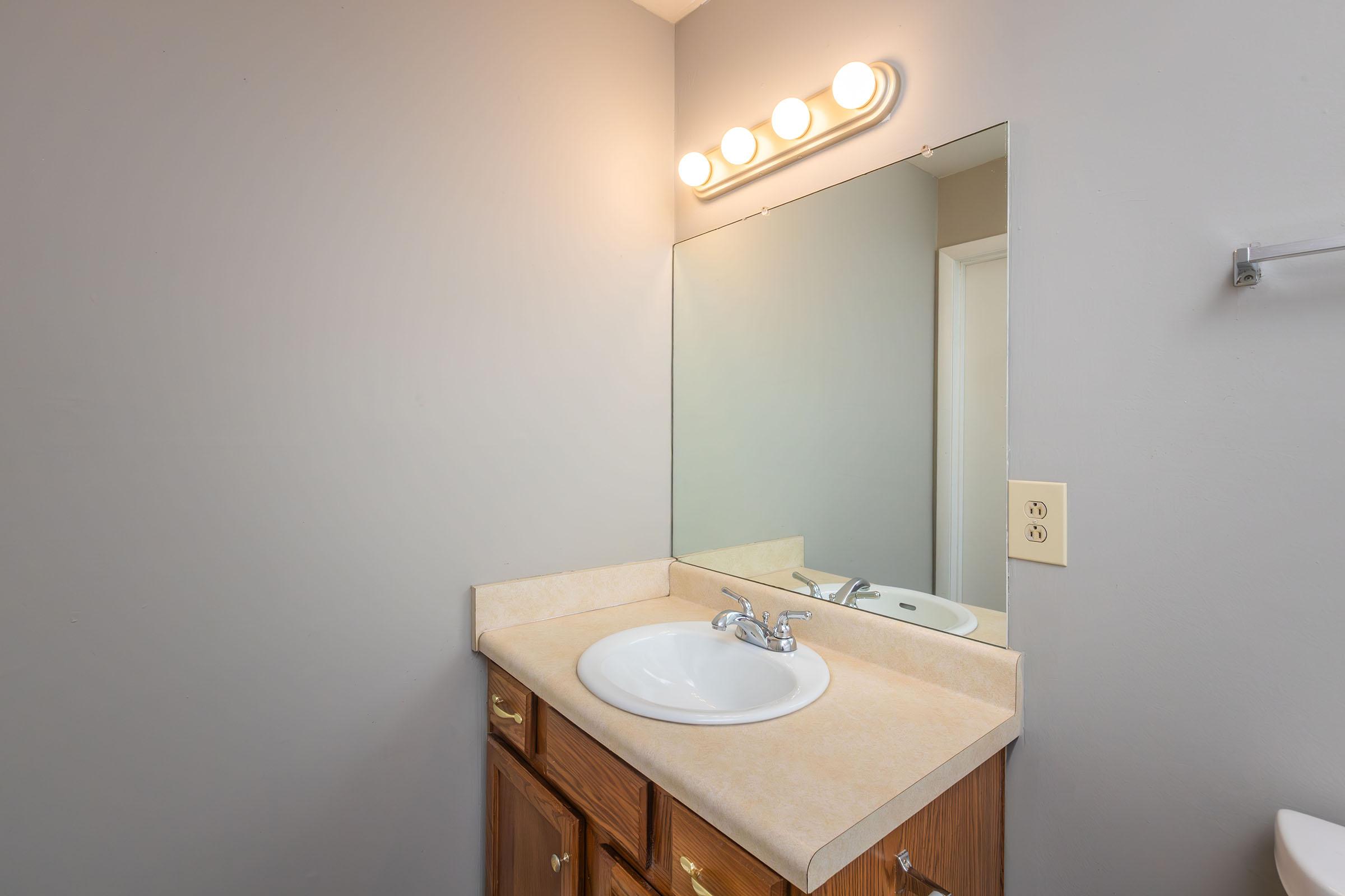 A clean, well-lit bathroom vanity with a light fixture above the mirror, a small round sink, and wooden cabinetry. The walls are painted in a neutral color, and the counter has a simple design. A towel rack is visible on the side, and the scene conveys a tidy and functional bathroom space.