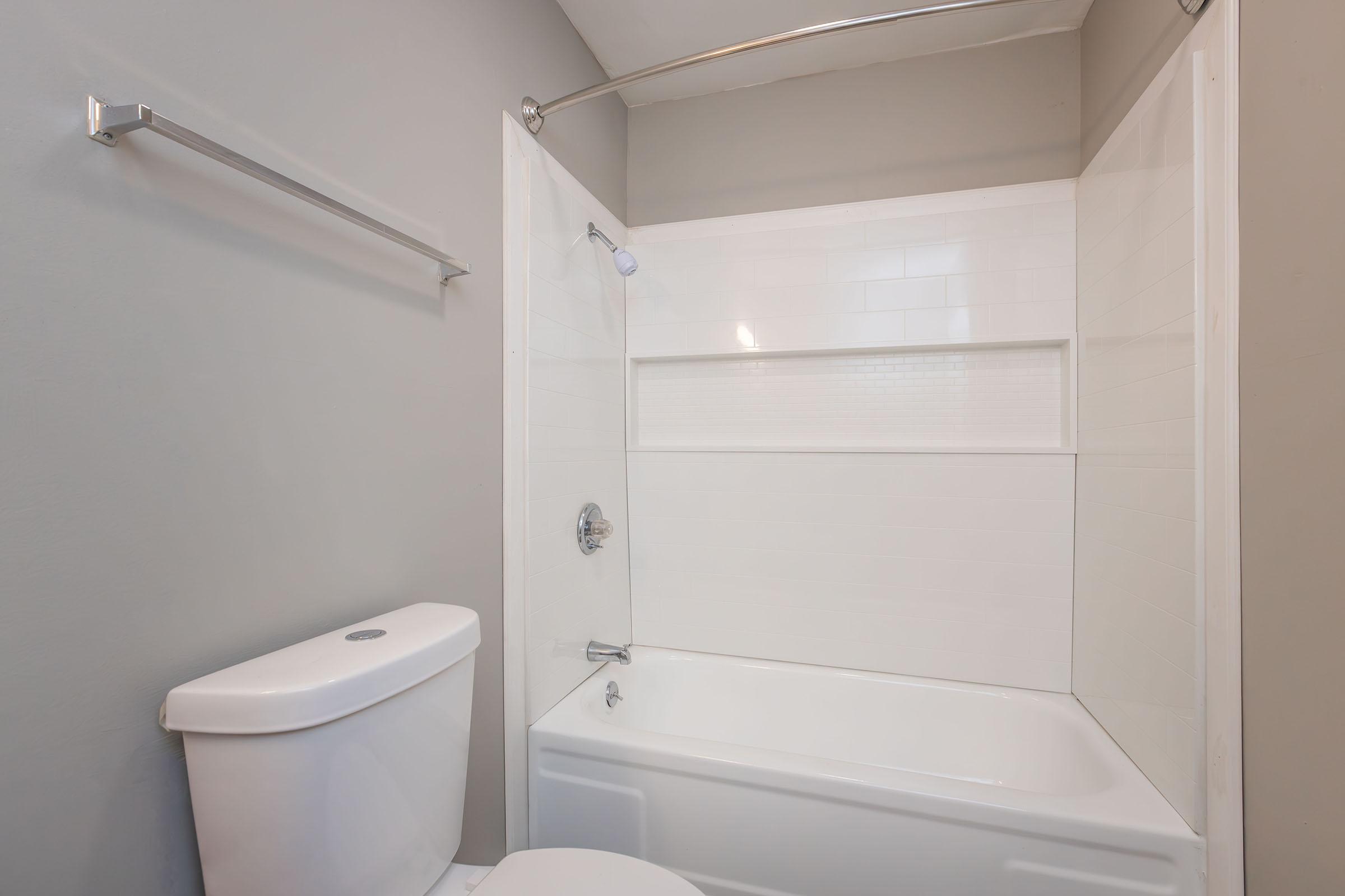 A clean and modern bathroom featuring a bathtub with a shower, a toilet, and light gray walls. The bathtub has a clear glass shower screen and a built-in shelf for toiletries. A towel rack is mounted on the wall beside the shower.