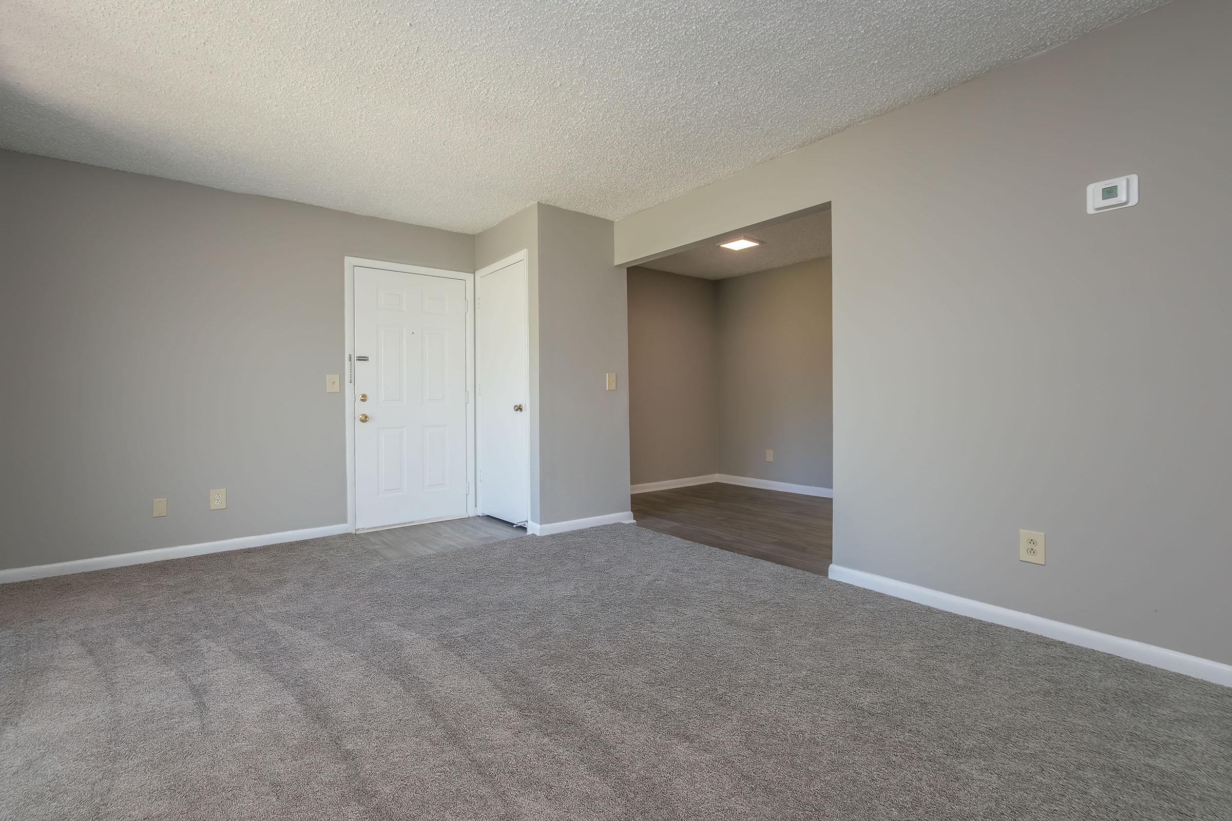A vacant interior space with light gray walls and carpeted flooring. The entrance features a white door, and there is a small opening leading to another room. Natural light enters from a nearby window, creating a bright and airy atmosphere. The layout is simple and uncluttered, suitable for various uses.
