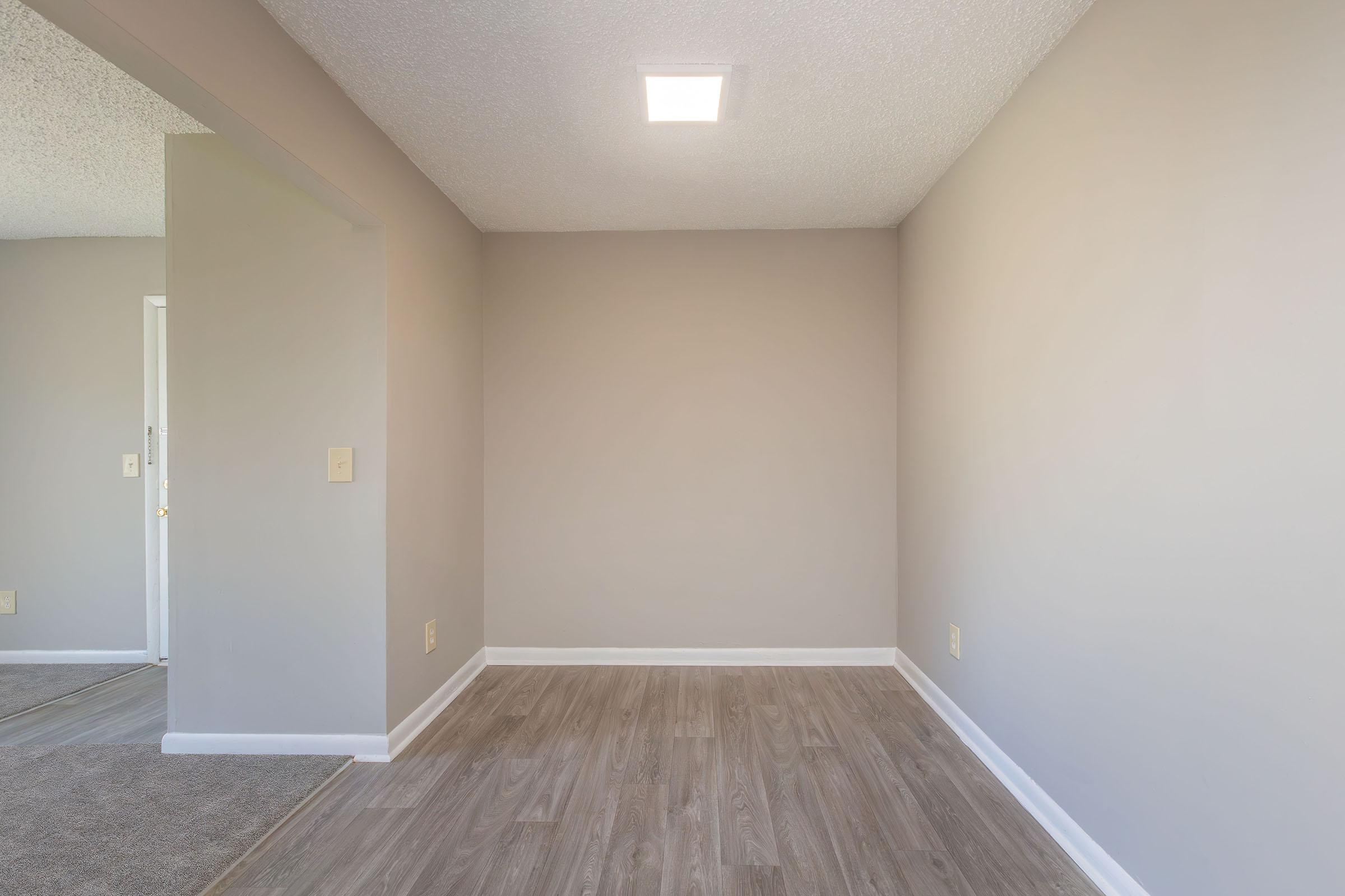 A simple, empty room with light gray walls and a modern ceiling light. The floor is covered in light wood laminate. An unfinished corner leads to another part of the space. The overall atmosphere is bright and minimalistic, creating a blank canvas for decoration or furniture.