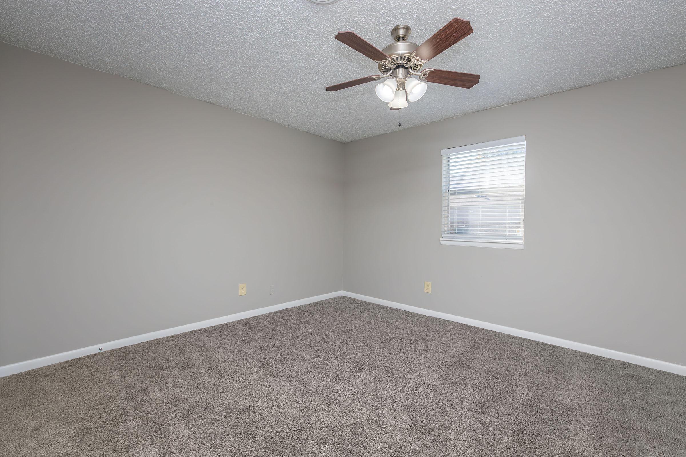 Empty room with light gray walls and a ceiling fan. The floor is covered with brown carpet, and there is a small window with white blinds allowing natural light in. The overall space is unadorned and clean, suitable for use as a bedroom or office.