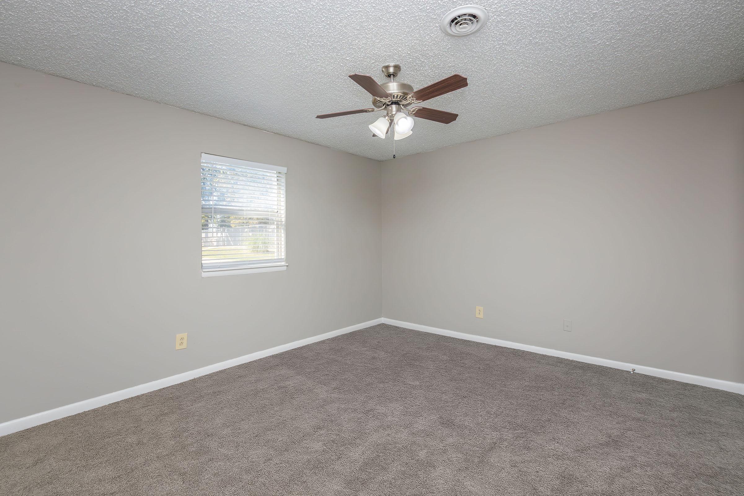 Empty room with light beige walls and a ceiling fan. The floor is covered with plush, light brown carpet. A single window with white blinds allows natural light to enter. The space appears clean and uncluttered, suitable for various purposes such as a bedroom or office.