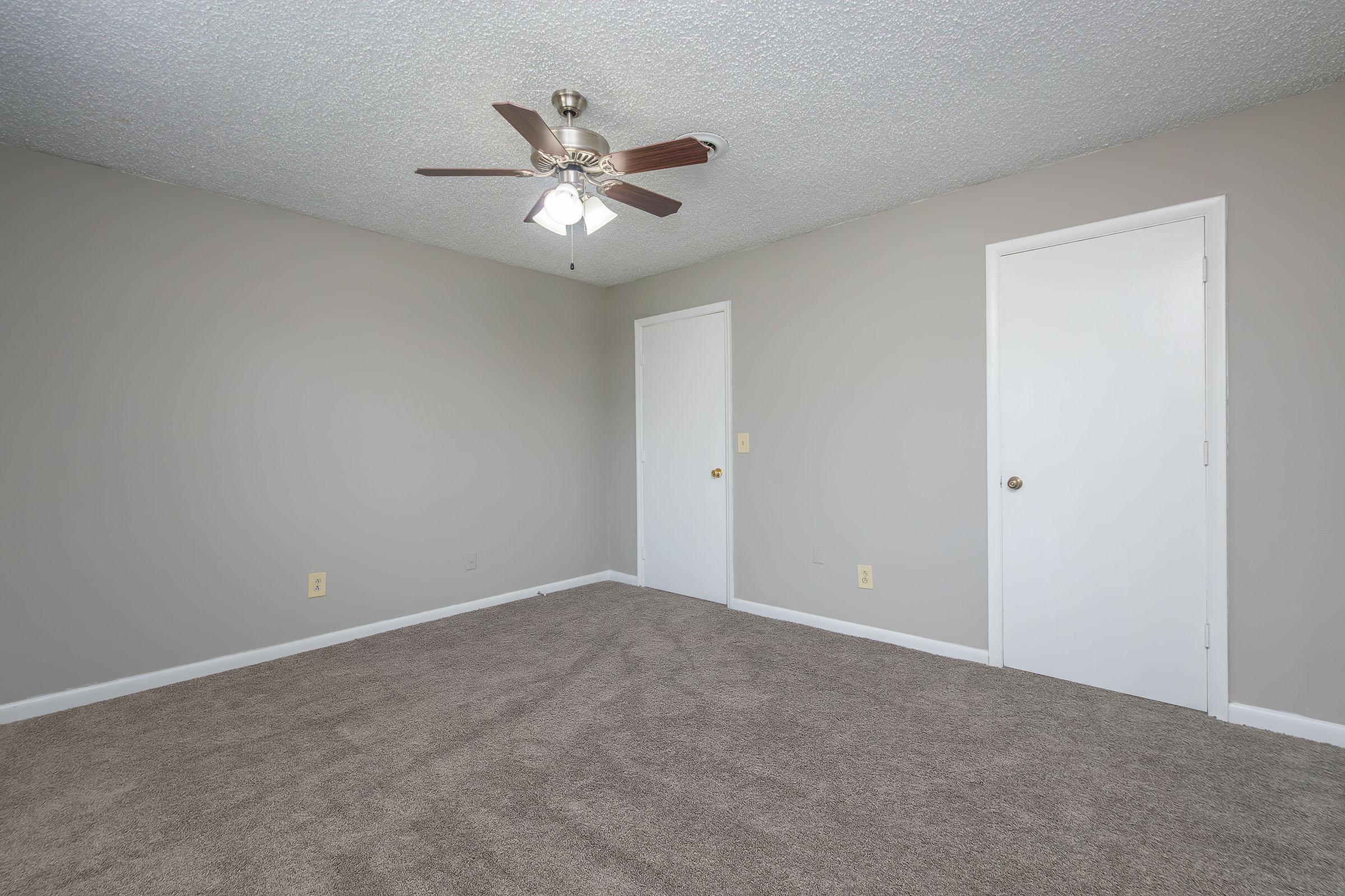 A spacious, empty room featuring light gray walls and freshly carpeted flooring. A ceiling fan with wooden blades is mounted in the center, and there are two closed white doors on opposite walls. The overall atmosphere is clean and ready for furnishing.