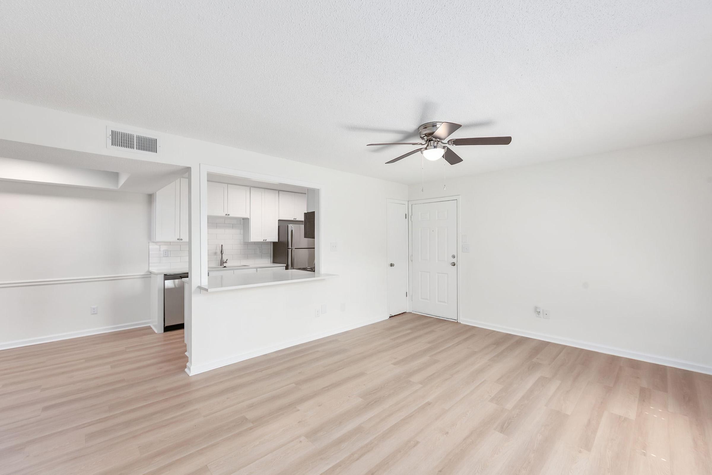 Bright and spacious living area with light-colored walls and hardwood flooring. A ceiling fan is mounted on the ceiling. An opening leads to a modern kitchen featuring white cabinetry and appliances. The entrance door is visible, and there are large windows allowing natural light to fill the space.