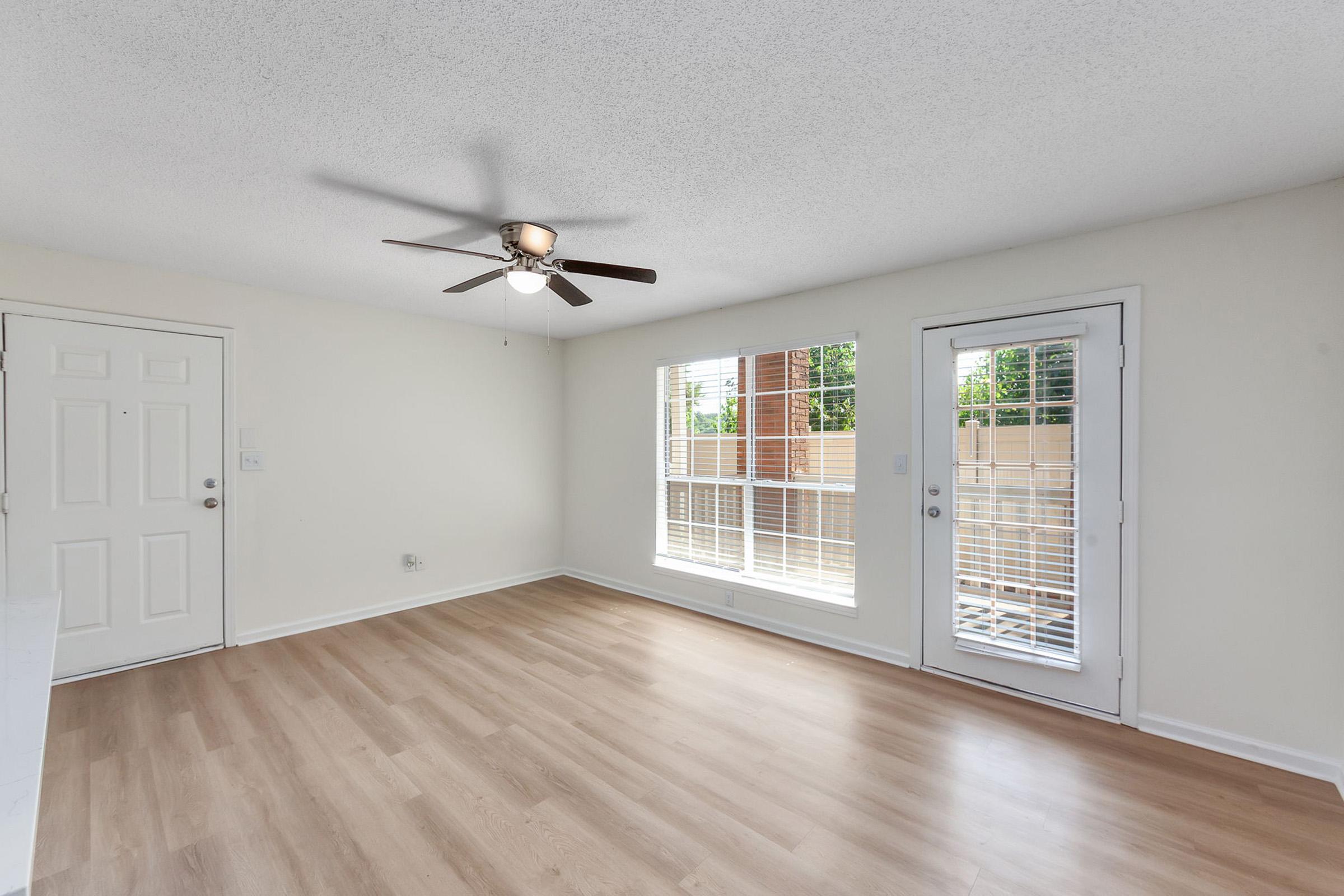 A well-lit living room featuring light-colored walls and a ceiling fan. The space has large windows with white blinds, and an entrance door leading outside. The floor is covered with light wood laminate, creating a warm and inviting atmosphere.