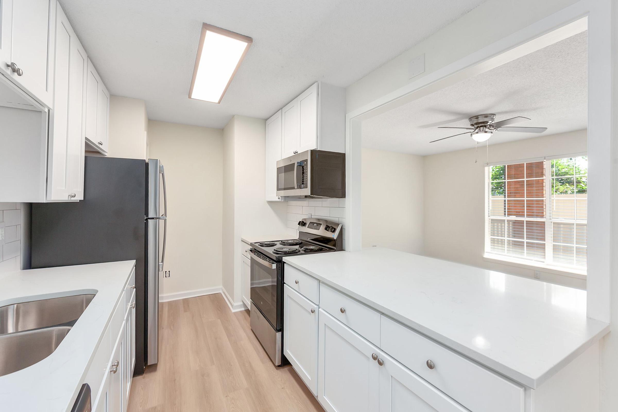 Modern kitchen featuring white cabinets, stainless steel appliances, and a light-colored countertop. A ceiling fan is visible in the adjacent dining area with large windows allowing natural light. The floor is light wood, creating a bright and open atmosphere.