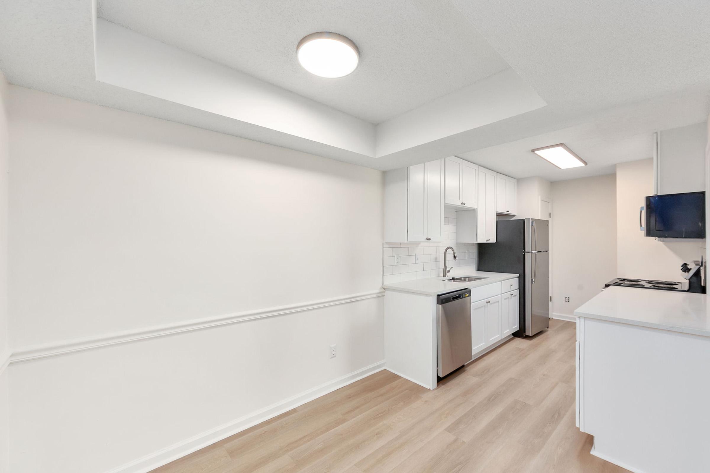 A modern kitchen featuring white cabinetry, stainless steel appliances, and a light wood floor. The layout includes a refrigerator, dishwasher, and a double-basin sink, with an abundance of natural light. The walls are painted in a neutral tone, creating a clean and spacious atmosphere.