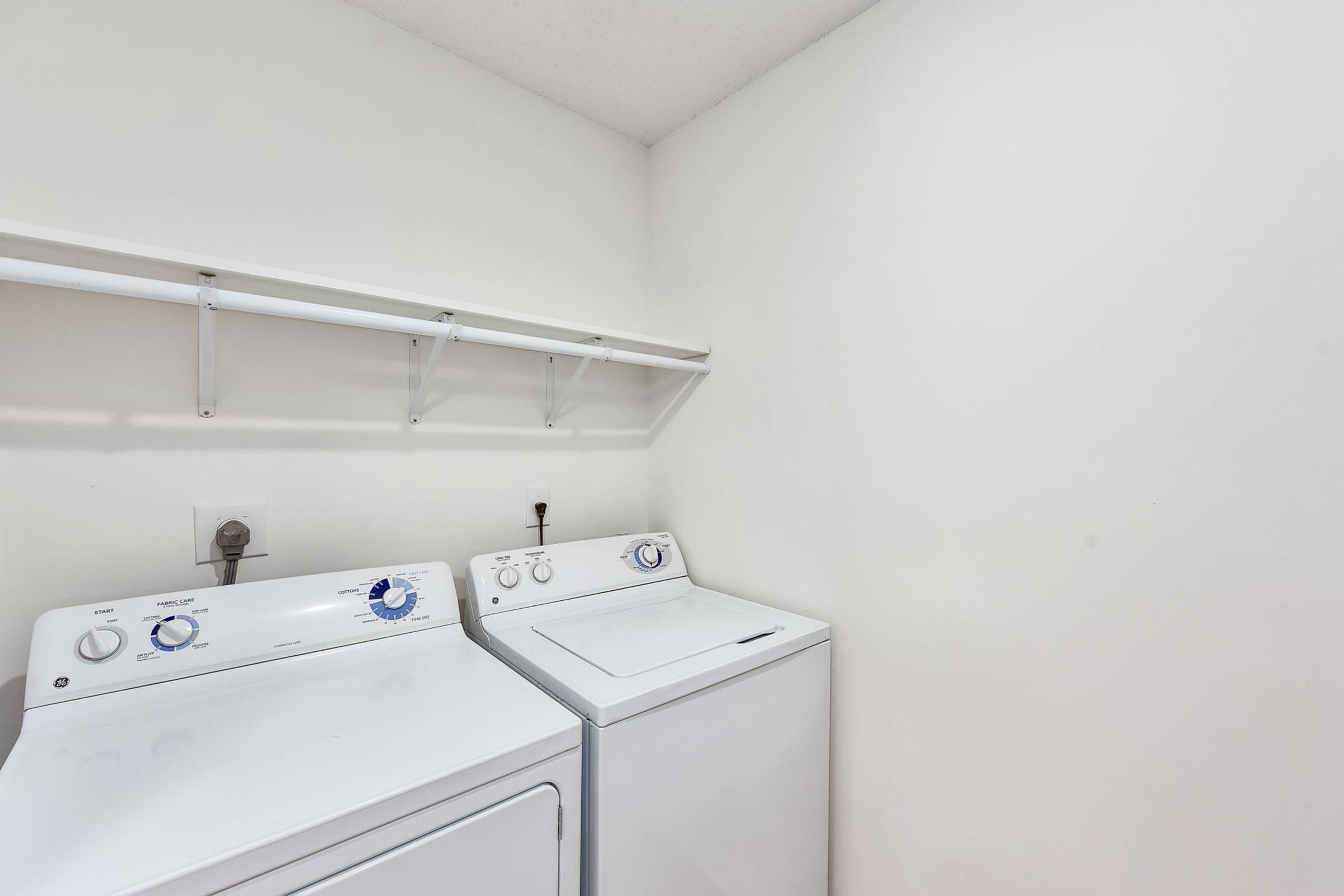 A clean, empty laundry room featuring a white washing machine and dryer side by side. There is a wall-mounted shelf above them for storage and the walls are painted a light color, creating a bright and spacious atmosphere.
