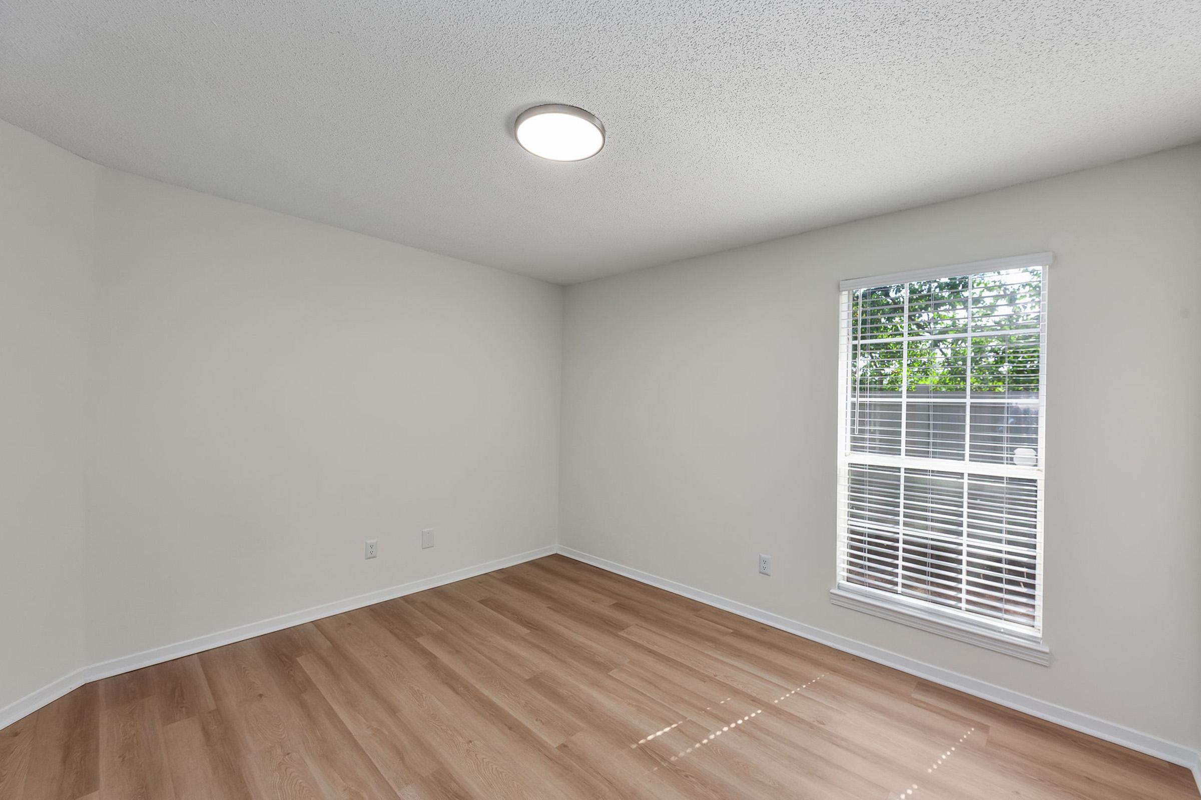 A clean, empty room with light-colored walls and a wooden floor. A round ceiling light provides illumination. A window with blinds allows natural light to enter, casting shadows on the floor. The space is minimalistic and uncluttered, ideal for various uses or personalization.