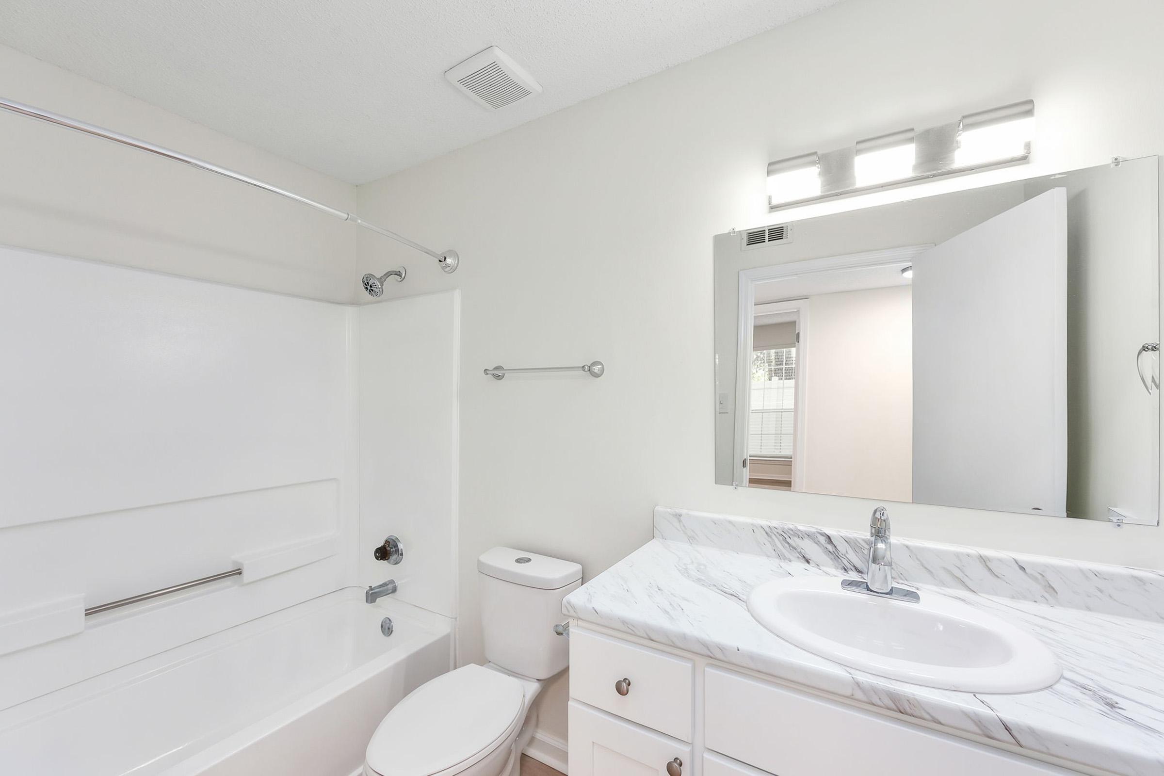 A clean and modern bathroom featuring a white bathtub with a shower, a marble countertop sink with a faucet, a toilet, and a wall mirror. The walls are painted in a light color, and there is ample lighting from above, creating a bright atmosphere. The door is partially open, revealing additional space.