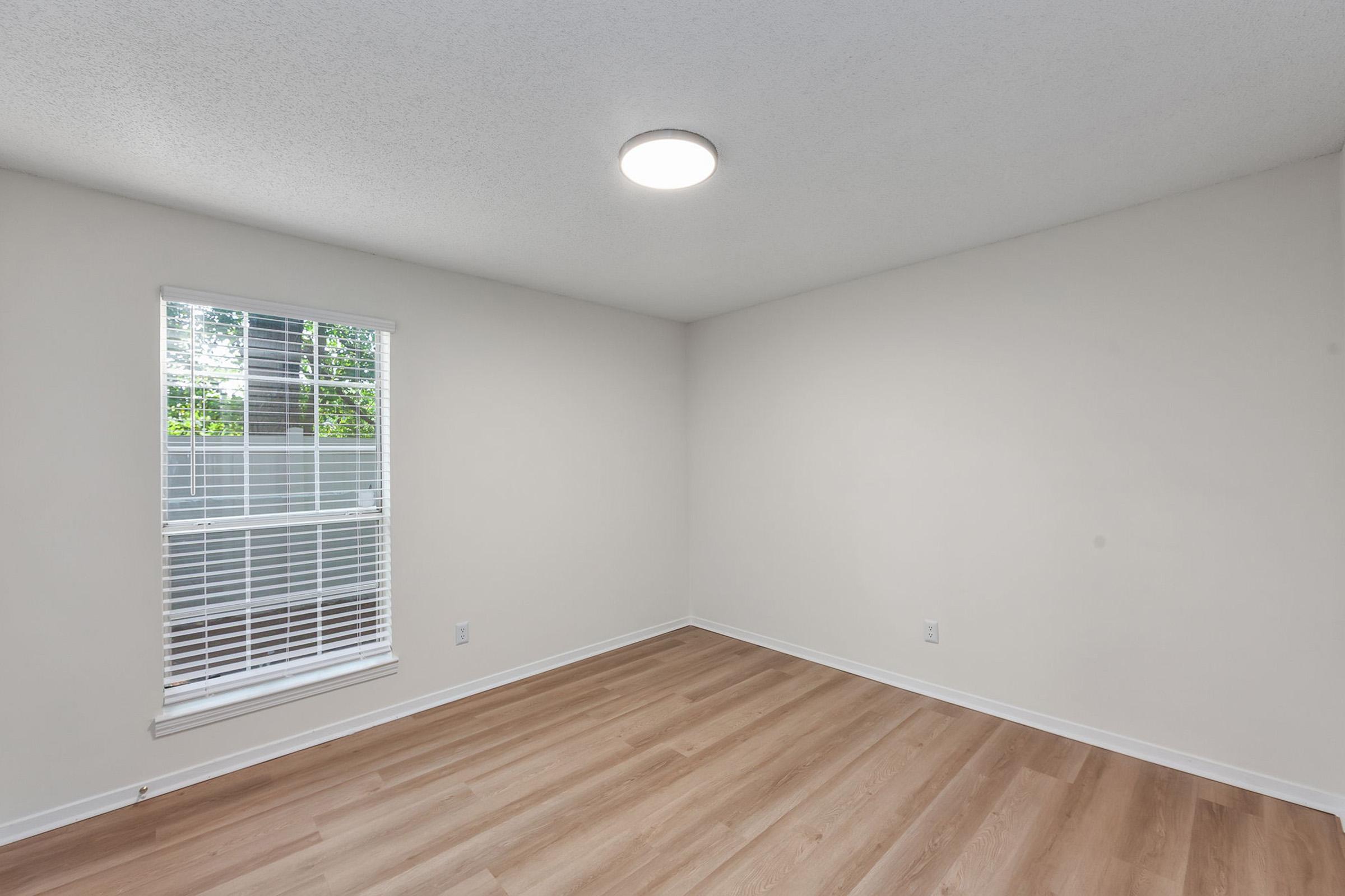 A spacious, empty room with light-colored walls and wooden flooring. A single window with white blinds allows natural light to stream in, and a modern ceiling light fixture illuminates the space. The room appears clean and ready for furnishing.
