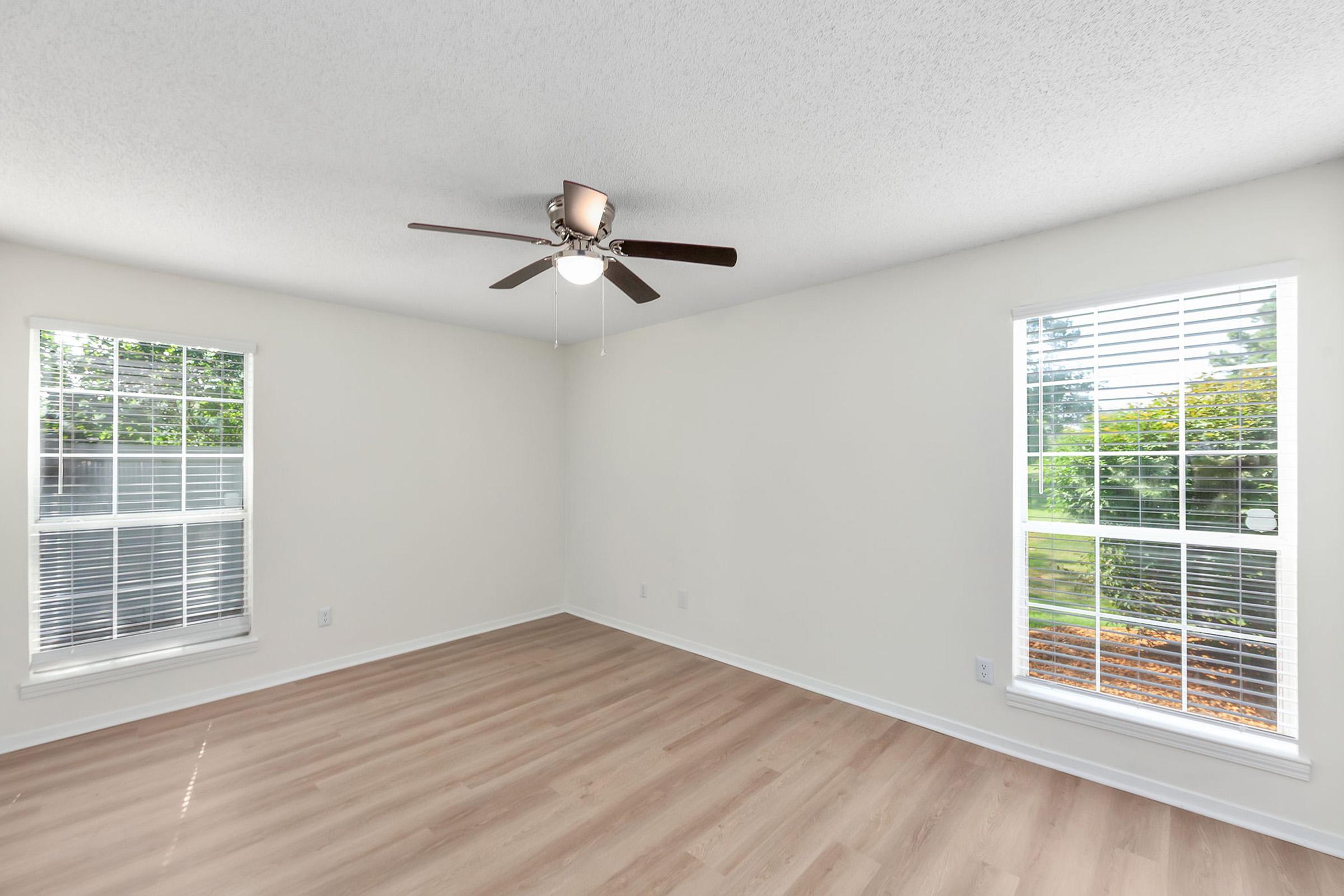 A bright, empty room with light-colored walls and a light wood floor. Two windows with white blinds allow natural light to enter. A ceiling fan with wooden blades is centered in the room, adding a modern touch. The surroundings outside are visible through the windows, showcasing greenery.