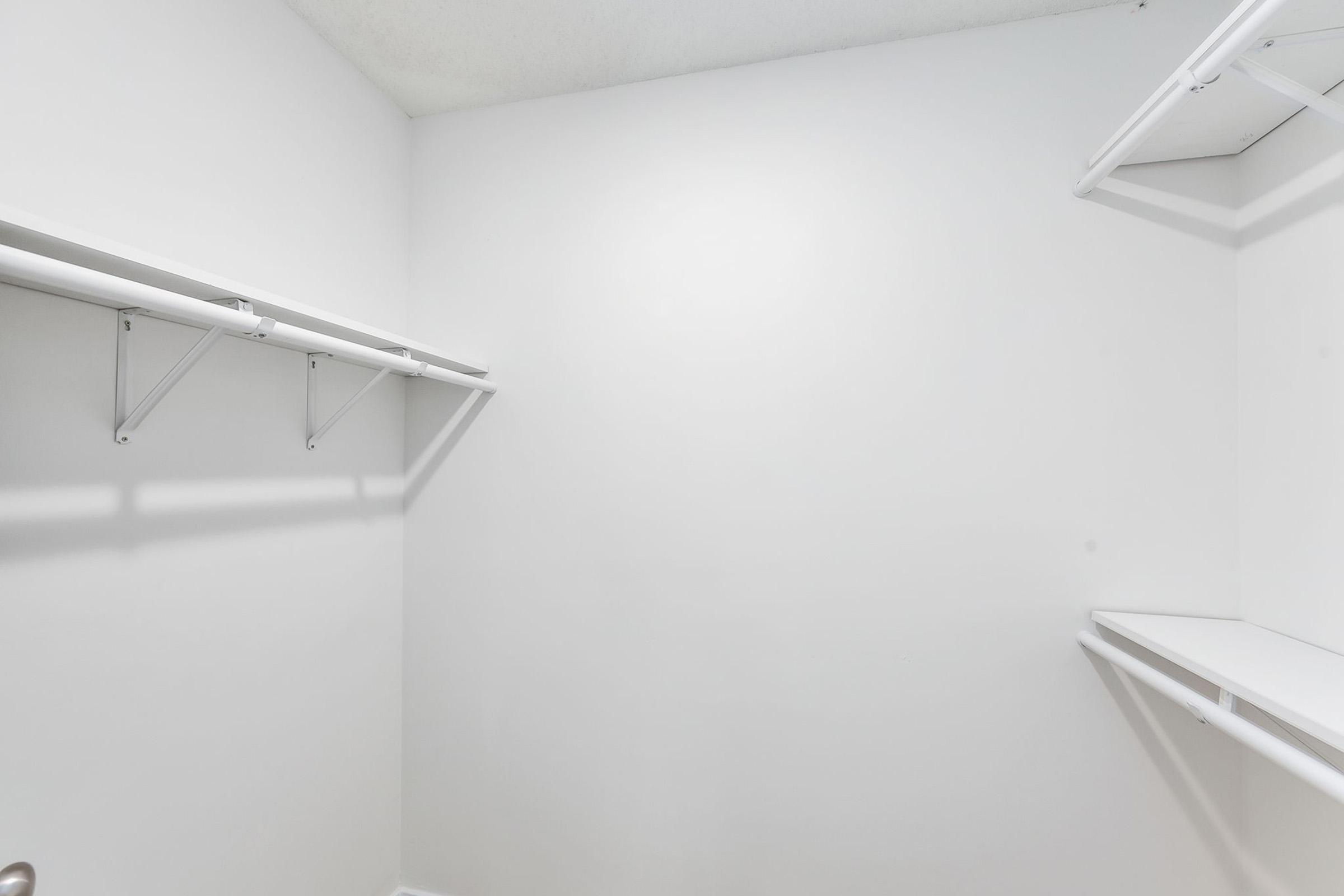 Empty, minimalist closet with bare white walls and no shelves or stored items. There are two horizontal railings on opposite walls for hanging clothes, but the space appears unorganized and unused, conveying a sense of emptiness and simplicity.