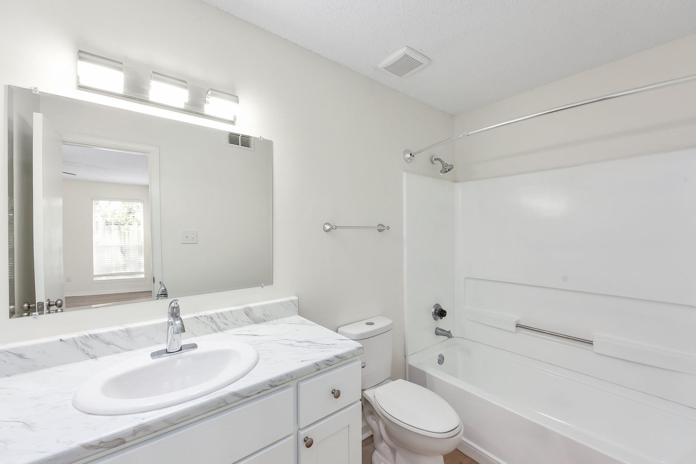 Modern bathroom featuring a marble countertop with a sink, a white toilet, and a bathtub with a shower. The space is well-lit with overhead lighting, and a large mirror enhances the room's brightness. Neutral-colored walls create a clean and simple aesthetic. Natural light filters in through a window.