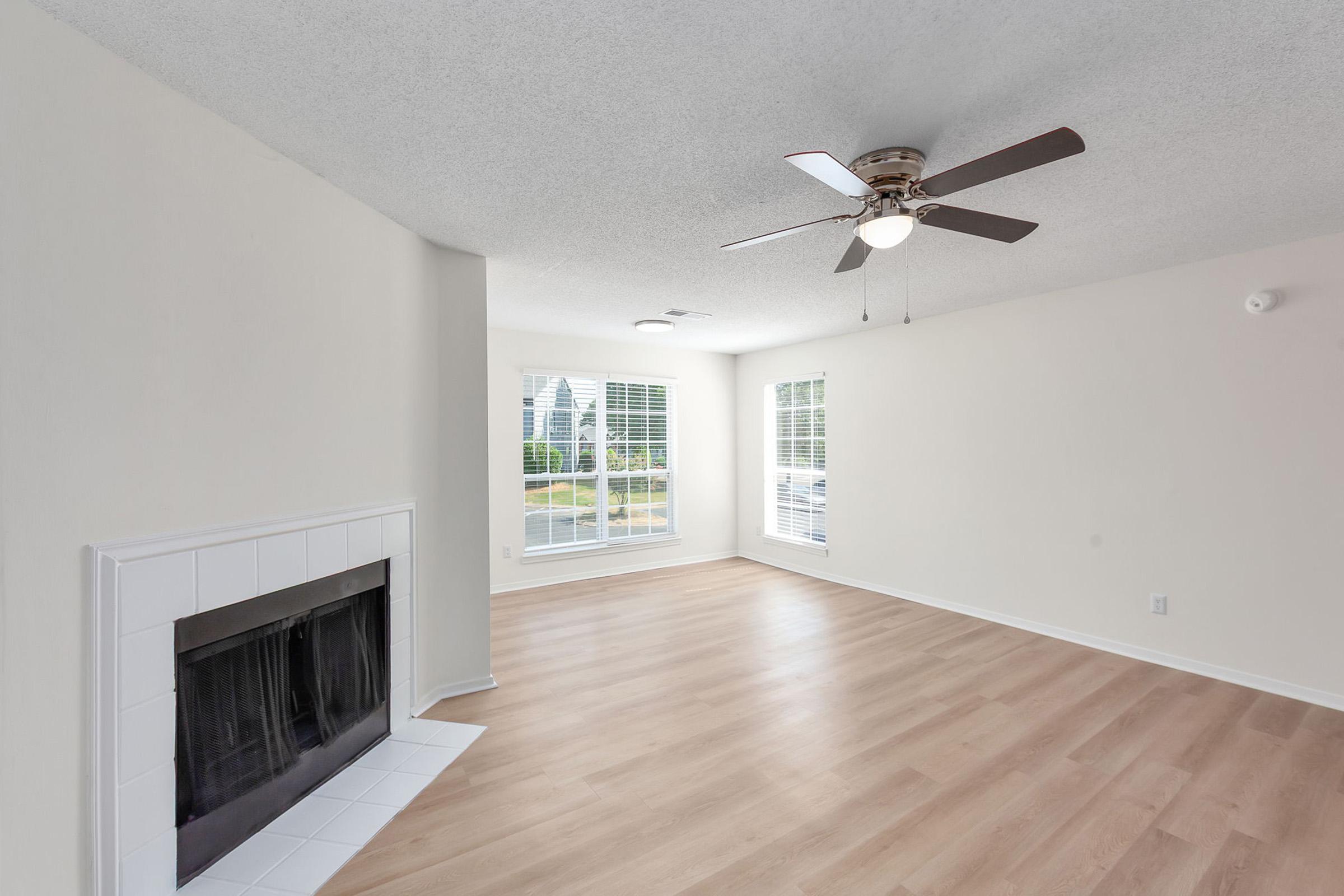 A bright, spacious living room featuring light-colored walls, a ceiling fan, and a large window with blinds. There is a white tile fireplace on one wall and light wooden flooring throughout the room. The area is uncluttered, creating an open and inviting atmosphere.
