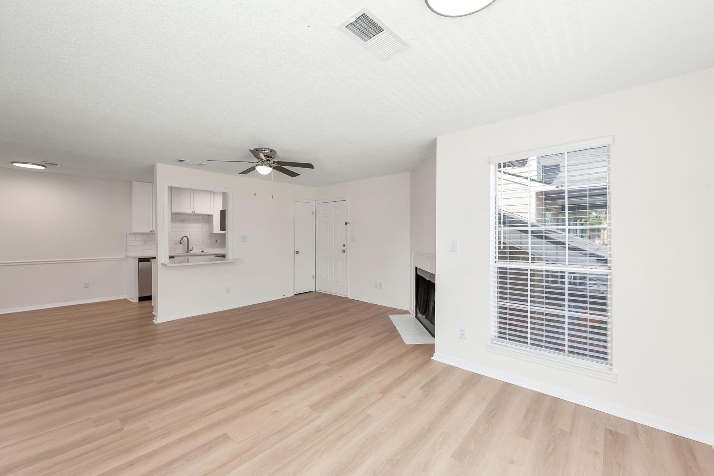 Spacious living area with light-colored walls and wood-style flooring. A ceiling fan is mounted above, and a window with blinds provides natural light. A doorway leads to a kitchen area, and there is an empty fireplace on one wall. The overall ambiance is bright and inviting.