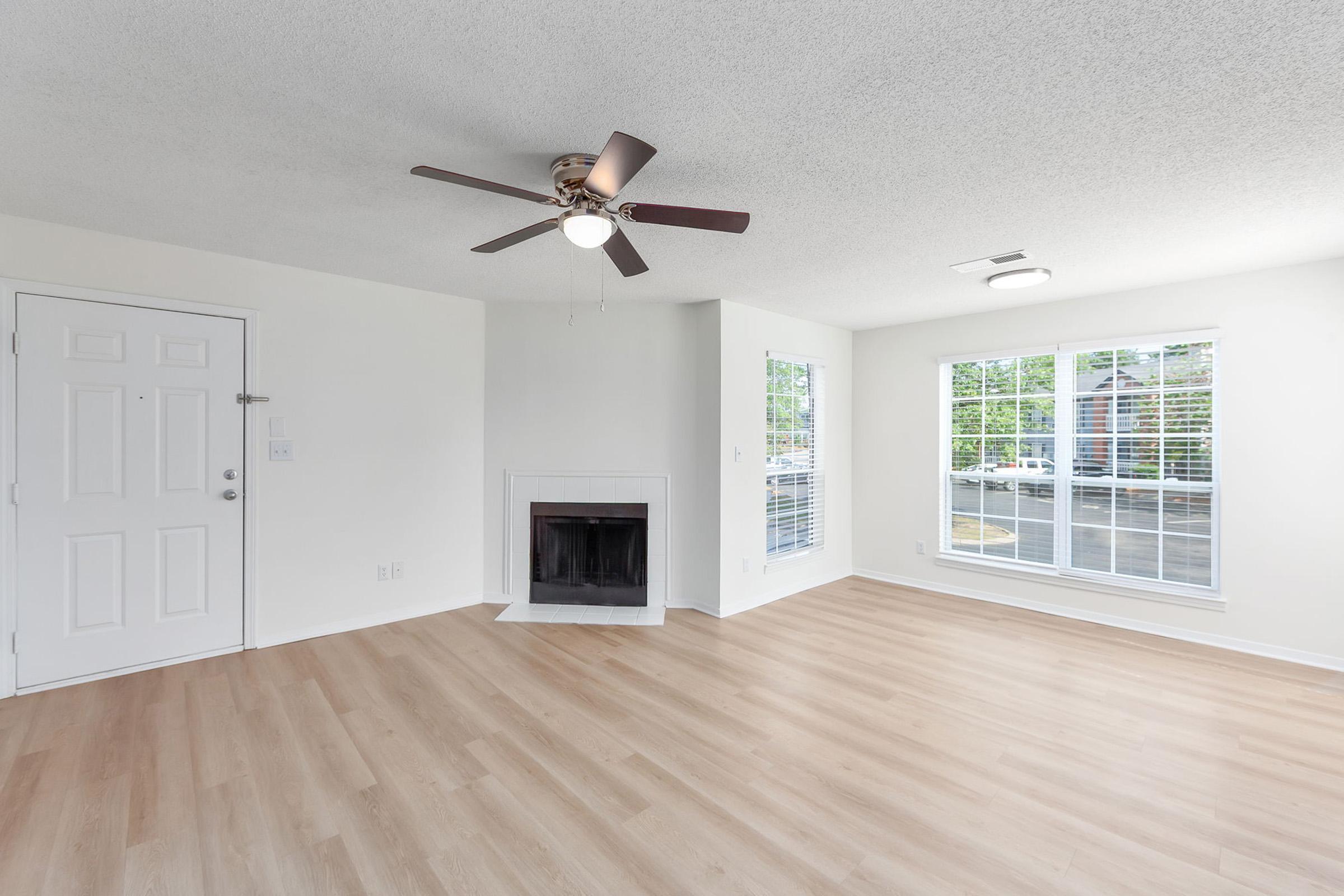 A spacious, well-lit living room featuring light wood flooring, white walls, and a ceiling fan. A cozy fireplace is located on one wall, and large windows provide natural light with views outside. The front door is visible to the left, along with a compact entryway.
