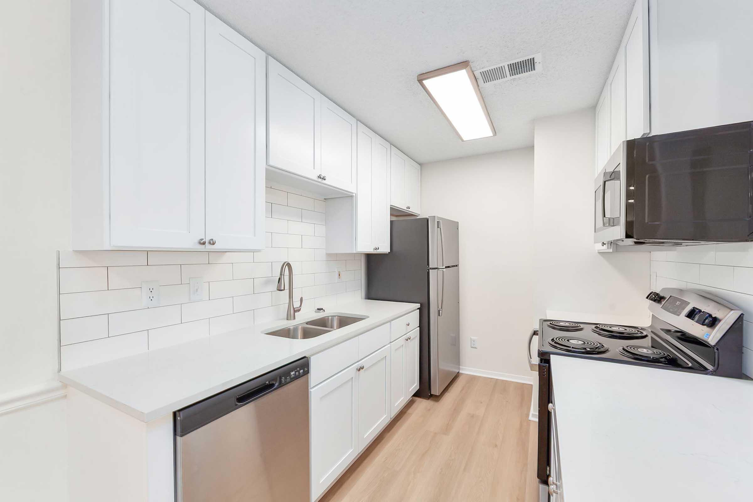 Modern kitchen featuring white cabinets, stainless steel appliances, a double sink, and a flat-top stove. The countertops are light-colored, and there's a tile backsplash. The flooring is a light wood laminate, creating a clean and bright atmosphere.