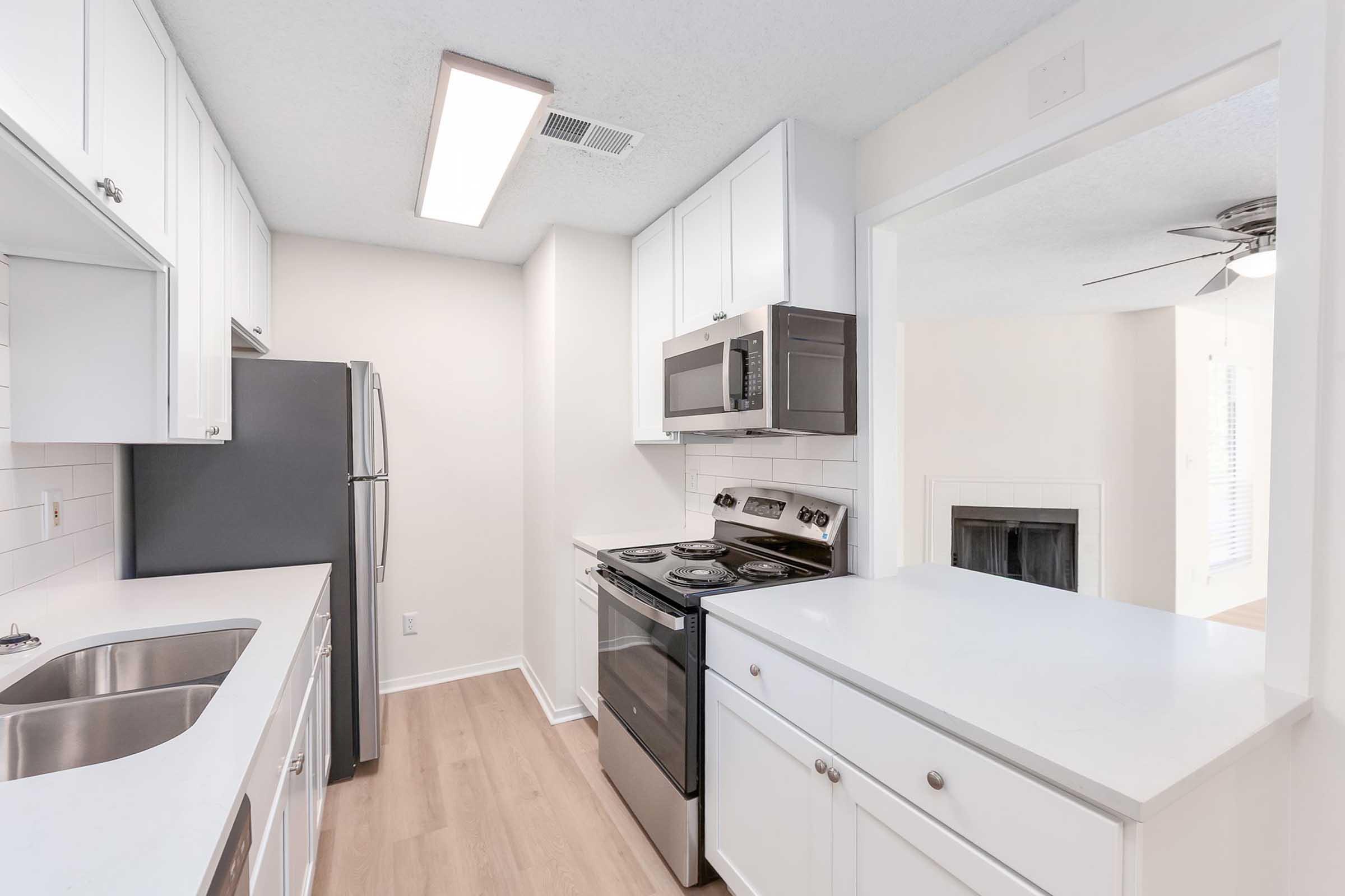 A modern kitchen featuring white cabinetry, stainless steel refrigerator, and black oven with a stovetop. The space has a bright, open layout with a pass-through counter and a clean, minimalist design, complemented by light wood flooring and natural light from a nearby window.