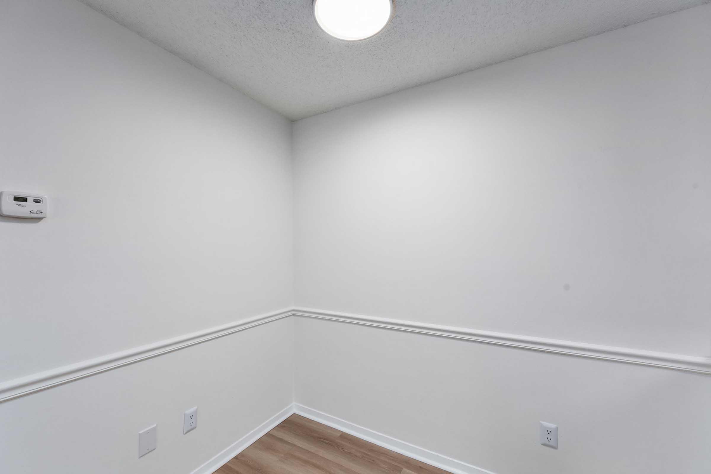 A corner of a room featuring white walls and a light ceiling. There is an overhead light fixture, a thermostat on the left wall, and a wooden floor. The room has a simple and clean design, with no furniture or decorations visible.