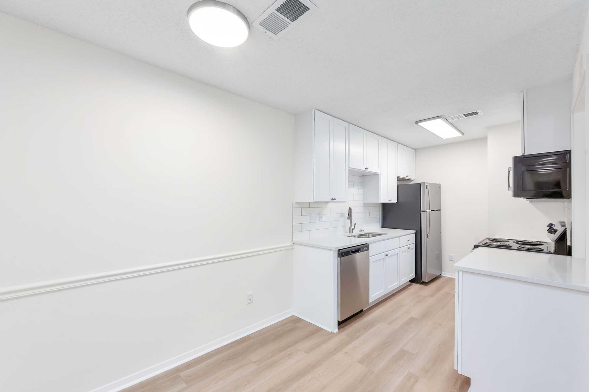 Modern kitchen featuring white cabinets, stainless steel appliances, including a refrigerator, dishwasher, and stove. The space has a light, neutral color scheme with laminate flooring and a clean, spacious layout. A ceiling light fixture provides overhead illumination.