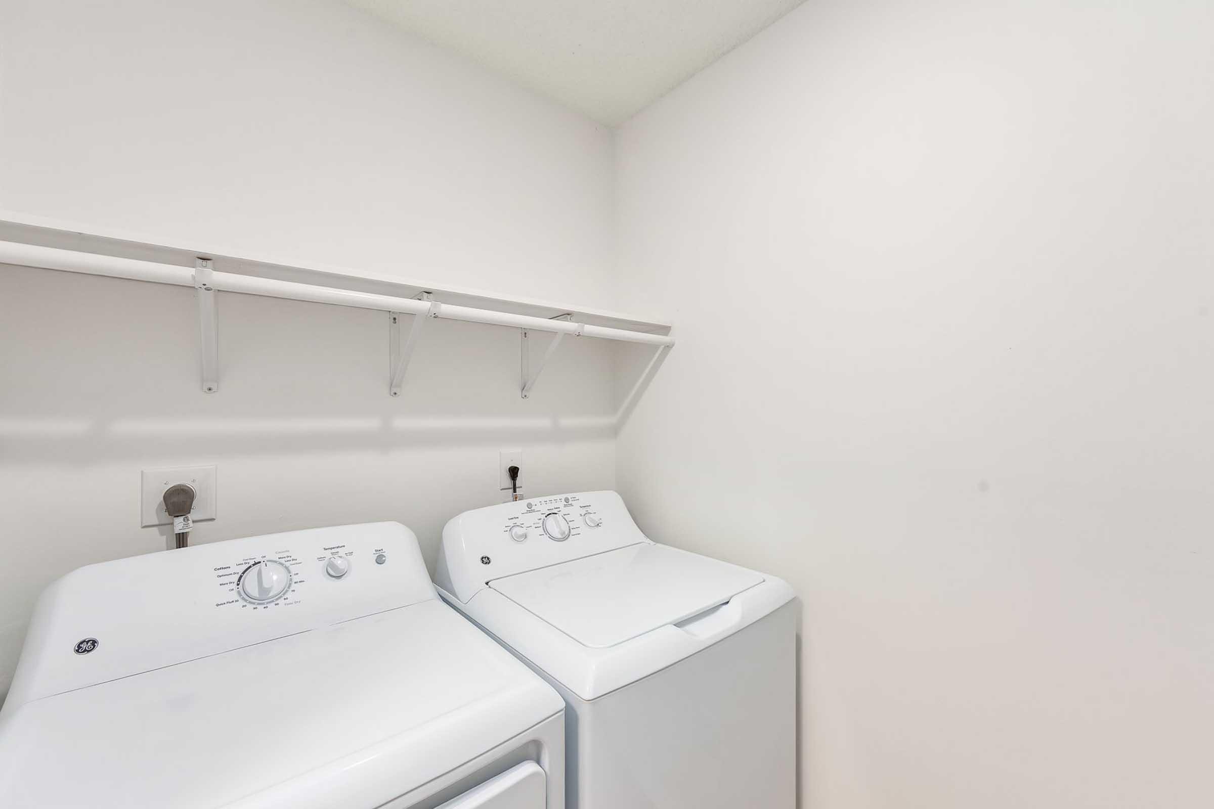 A clean and empty laundry room featuring two white washing machines side by side, with a simple wall-mounted shelf above them. The walls are plain and light-colored, contributing to a bright and uncluttered space.