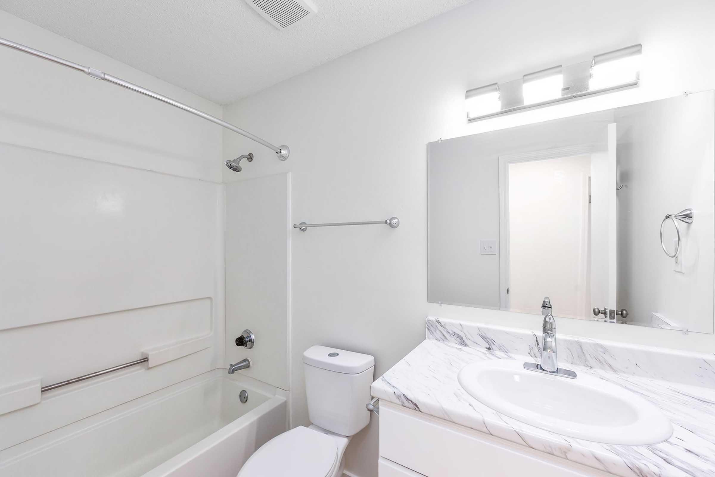 A clean and modern bathroom featuring a bathtub with a shower, a white toilet, and a sink with a marble countertop. The walls are painted white, and there is a mirror above the sink. Soft lighting is provided by a fixture above the mirror. The bathroom is well-lit and functional.