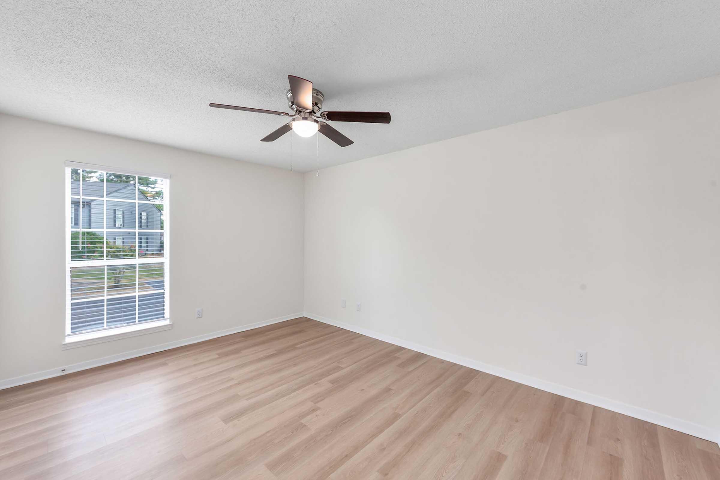 A bright, empty room featuring light-colored walls and a ceiling fan with wooden blades. There is a large window allowing natural light to enter, showcasing a view of the outdoors. The floor is covered in light wood-like laminate, enhancing the spacious feel of the area.