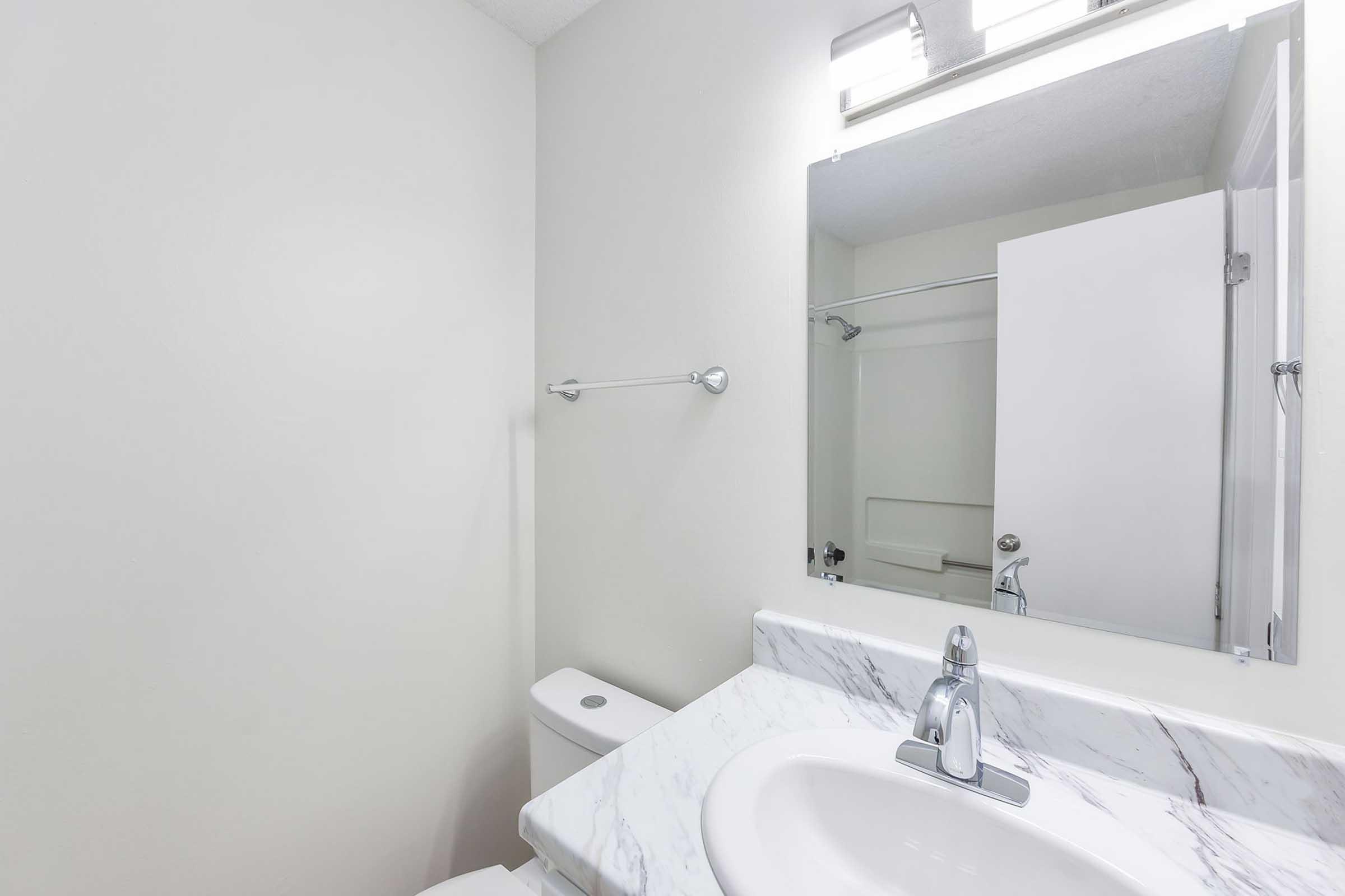 A clean, modern bathroom featuring a white sink with a marble countertop, a large mirror above it, and a toilet. A towel rack is mounted on the wall, and a white cabinet door is partially open, revealing a small shelf inside. The walls are painted a light color, enhancing the bright and airy feel of the space.