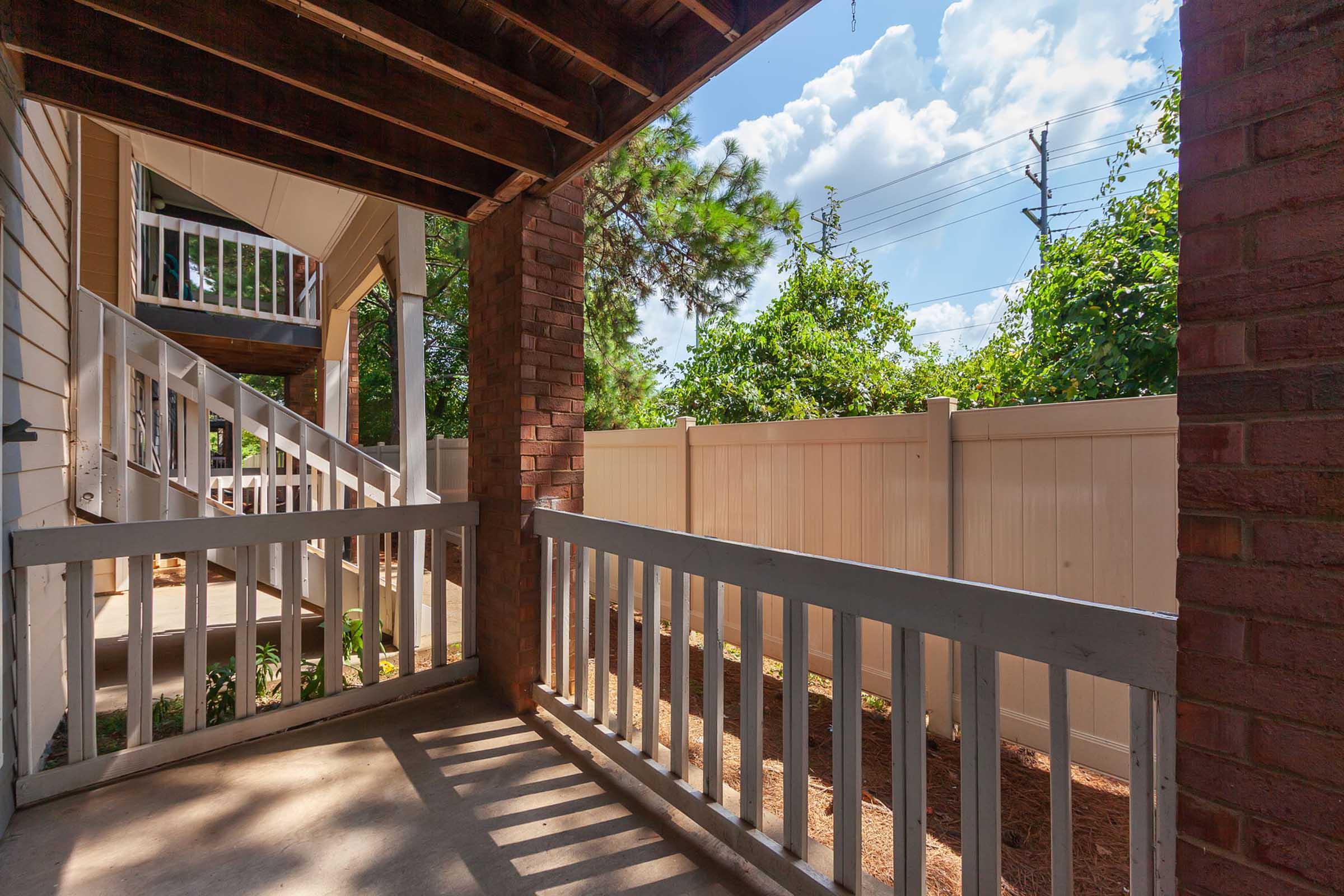 View of a porch with a wooden railing, looking out towards a garden area with greenery and a white fence. The sky is partially cloudy, with blue visible among the clouds. The surrounding area features a brick support column and a glimpse of additional steps leading to other levels of the building.