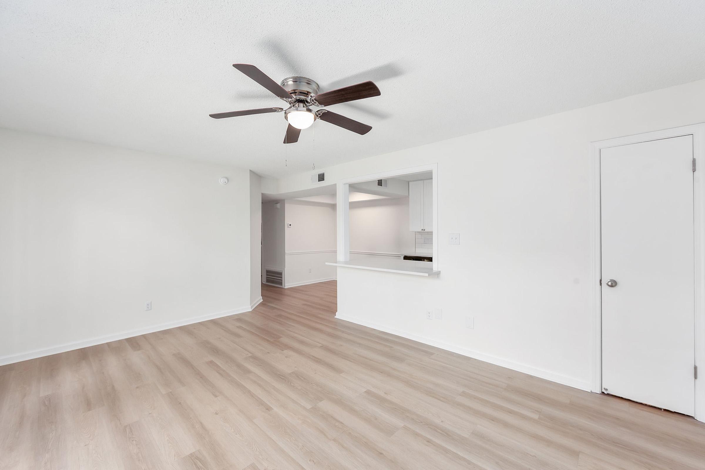 A bright, spacious room featuring light hardwood flooring, a ceiling fan with dark blades, and white walls. A pass-through window leads to an adjoining kitchen space, and a closed door suggests access to another room or area. The overall atmosphere is modern and inviting.