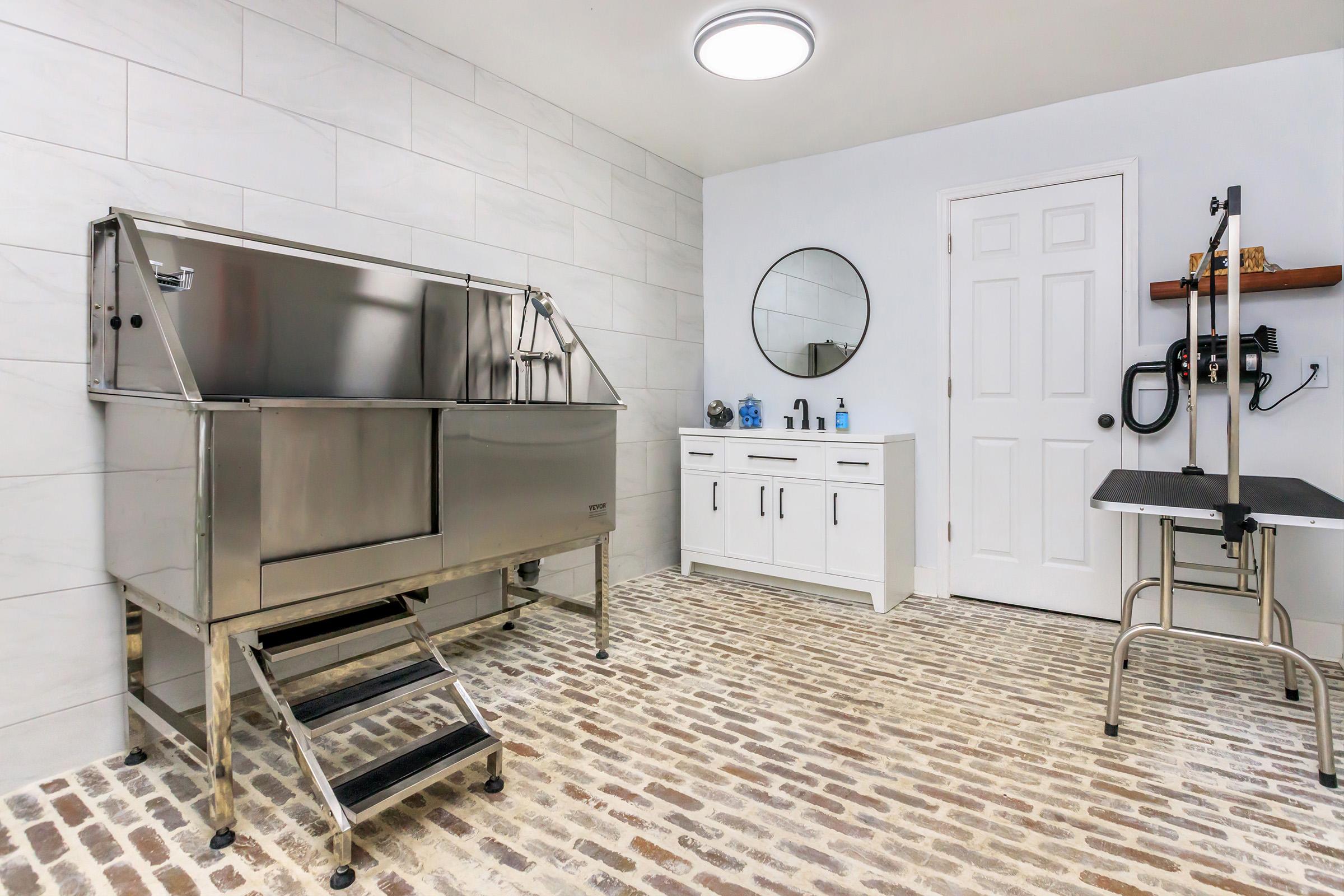 A spacious, modern pet grooming area featuring a stainless steel wash tub with steps, a round mirror, a white vanity with toiletries, and a grooming table. The floor is made of brick, and the walls are painted light gray, creating a clean and inviting atmosphere for pet care.