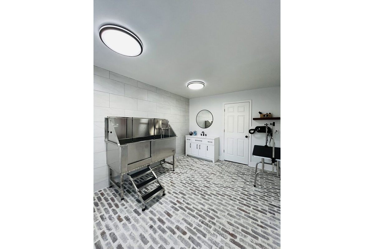 A modern grooming room featuring a stainless steel grooming table with steps, a circular mirror above a white vanity, and a door with a doorknob. The flooring consists of a cobblestone pattern, and there are ceiling lights providing bright illumination. The walls are tiled in a light color, creating a clean and spacious environment.