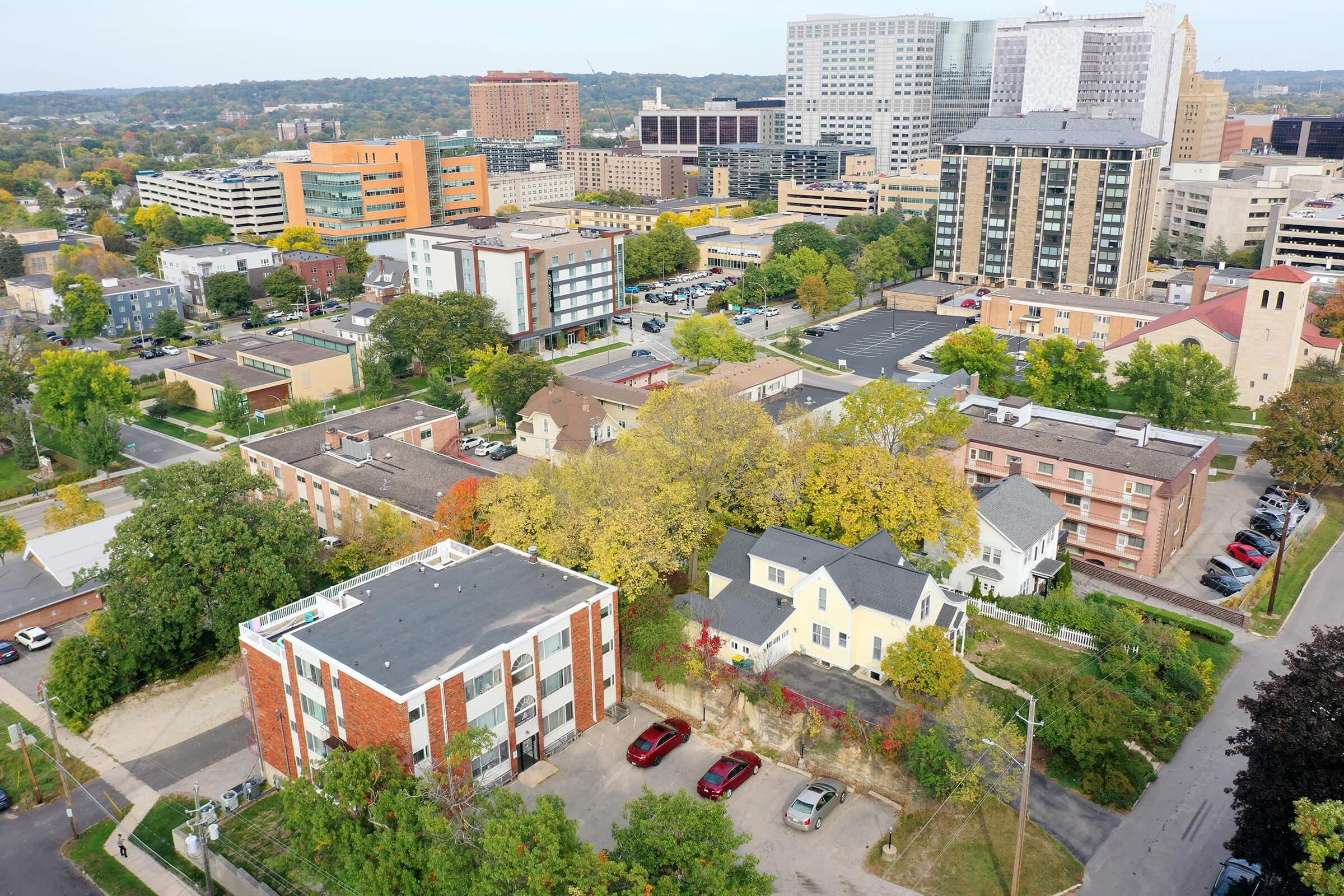 Aerial view of a cityscape showing a mix of residential and commercial buildings, green trees with fall foliage, and parked cars. The skyline features modern skyscrapers and low-rise buildings with various architectural designs, set against a background of hills and urban development.