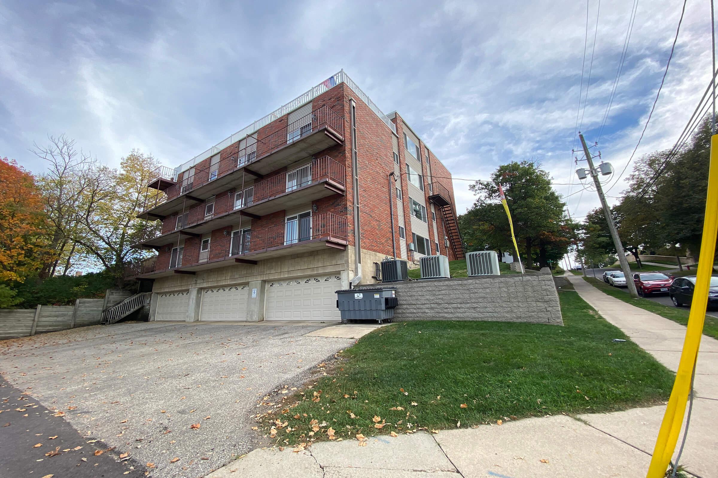 A three-story brick apartment building with several balconies and a garage area. The building is set back from the street, with grassy areas and a few autumn trees nearby. Utility poles and a sidewalk are visible, along with parked cars on the street. The sky is partly cloudy.