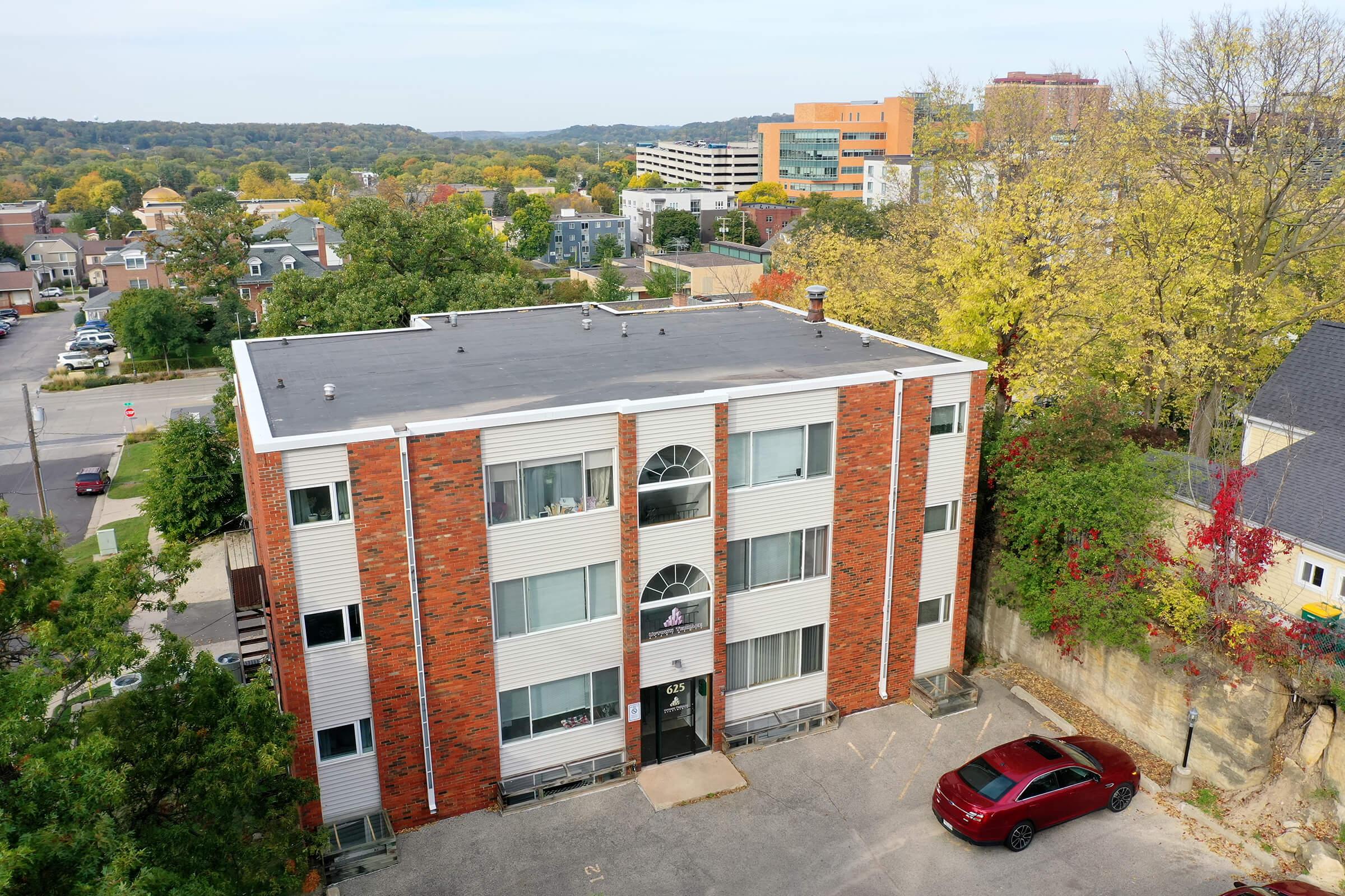Aerial view of a three-story brick apartment building with large windows, surrounded by trees and smaller buildings. A maroon car is parked in front, and the landscape includes a mix of urban and green areas, with a distant view of other structures and hills in the background.