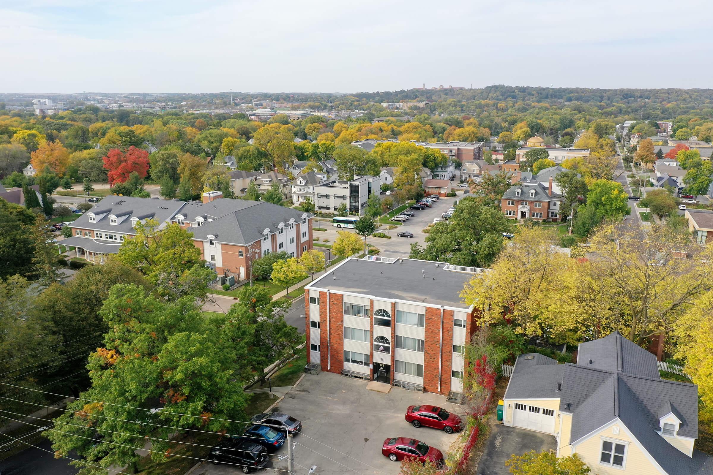 Aerial view of a suburban area featuring several buildings, including an apartment complex with parking, surrounded by trees displaying autumn colors. In the background, more buildings and greenery can be seen, showcasing a mix of urban and natural elements under a clear sky.