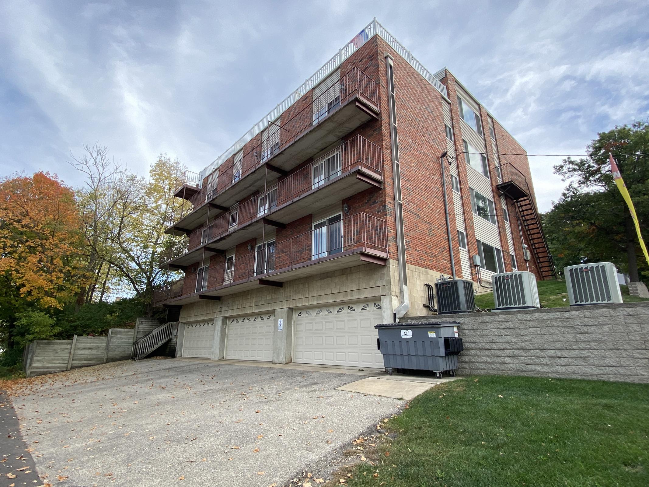 A multi-story brick building with balconies, set on a grassy area. Several air conditioning units are visible at the base, along with a row of garages. Fall foliage adds color to the scene, with trees displaying autumn leaves. The sky is partly cloudy, and a pathway leads up to the building.