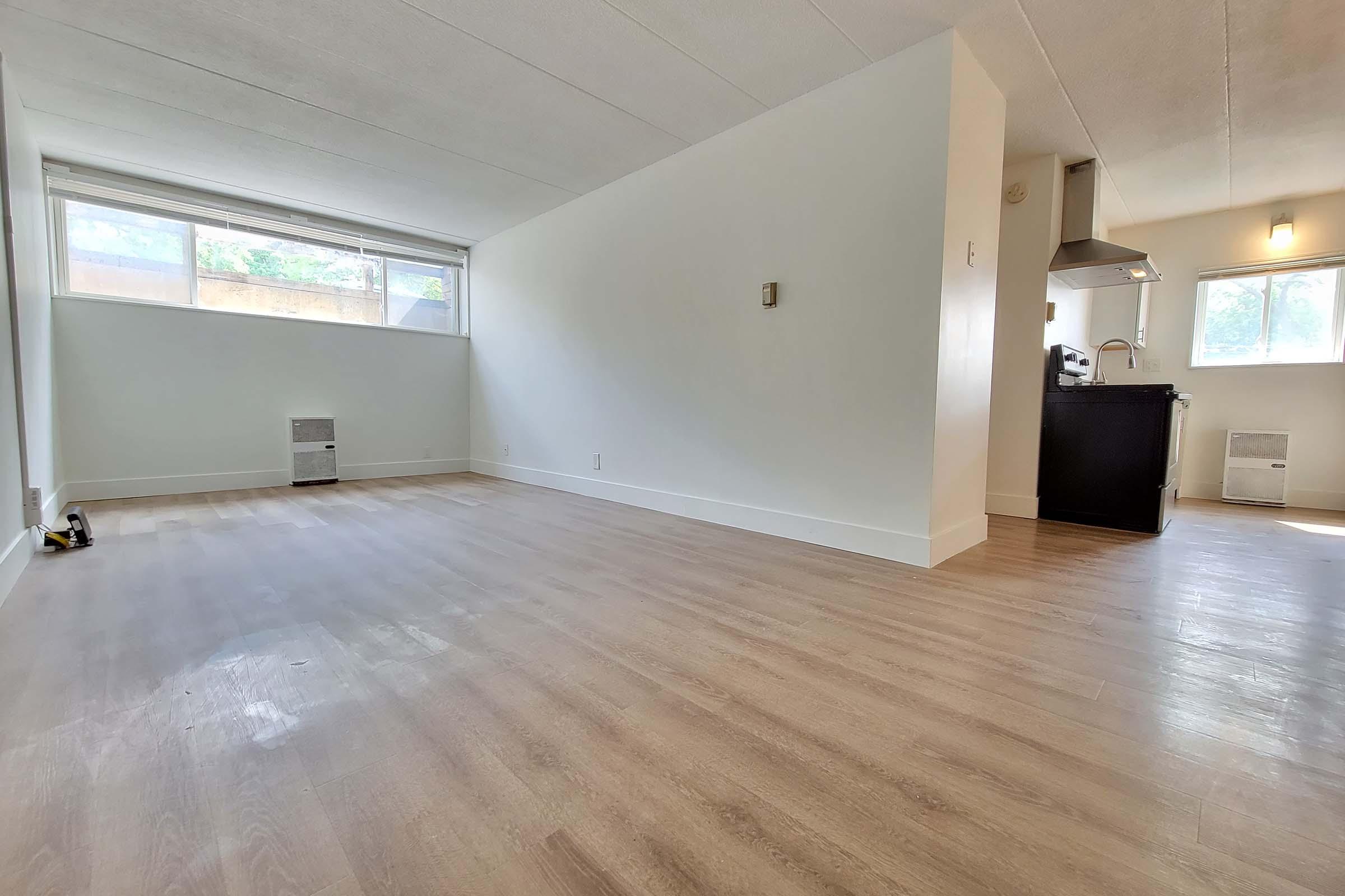 A spacious, empty room featuring light-colored wooden flooring, large windows allowing natural light, and a kitchenette in the background. The walls are painted white, creating a bright and airy atmosphere, with minimal furnishings.