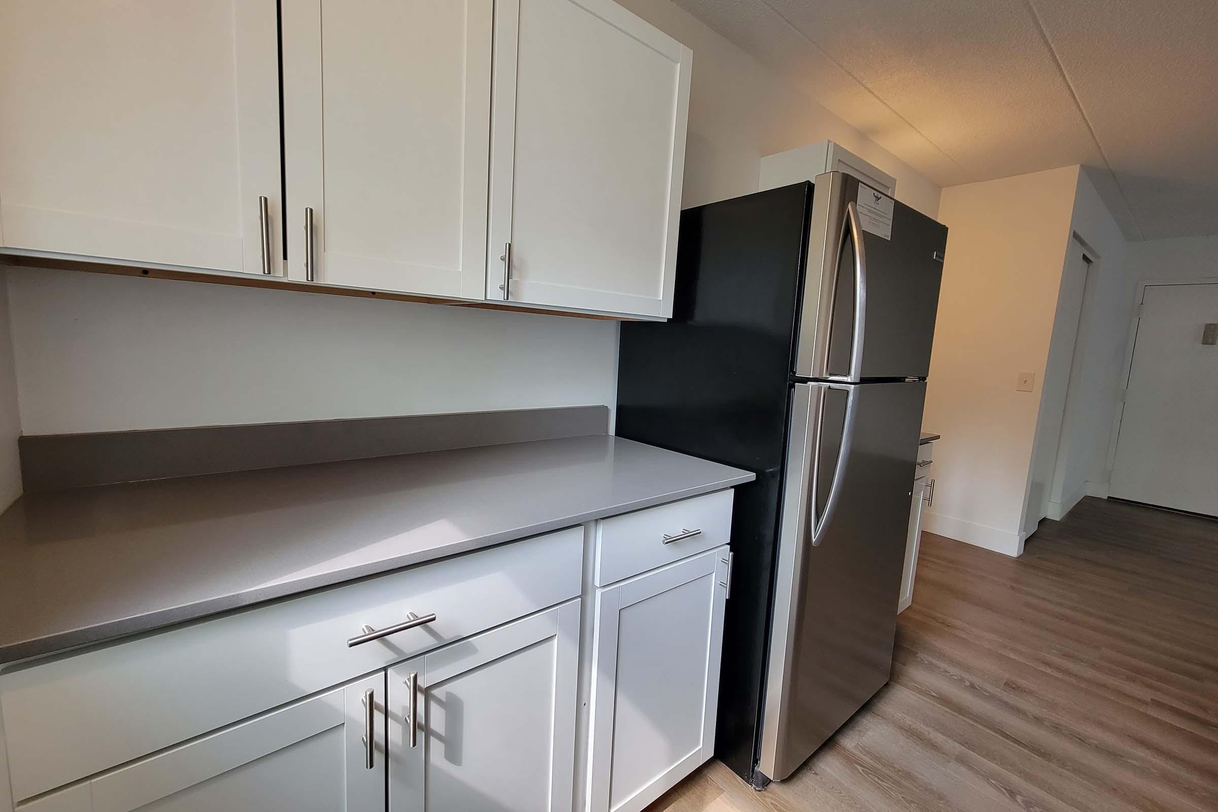 A modern kitchen with white cabinets, gray countertops, and a stainless steel refrigerator. The space is well-lit, featuring a clean and minimalist design with a light-colored floor and an open layout leading to a hallway.