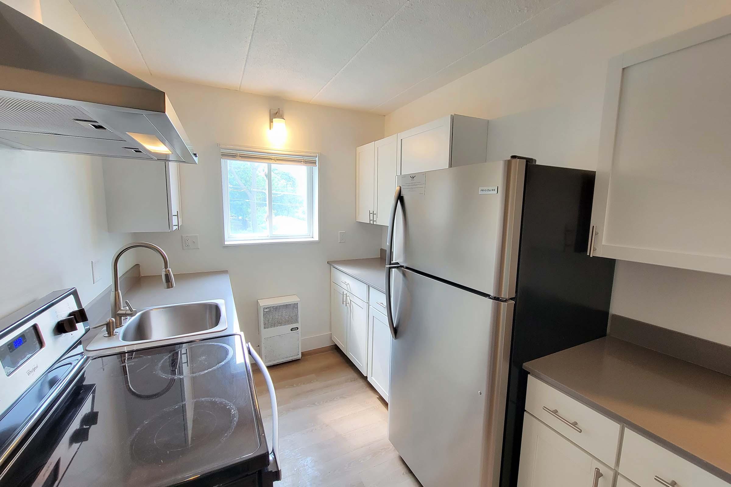 A modern kitchen featuring stainless steel appliances, including a refrigerator and stove. There are white cabinets and countertops, a stainless steel sink, and a window that lets in natural light. The flooring is light wood, creating a bright and inviting atmosphere.